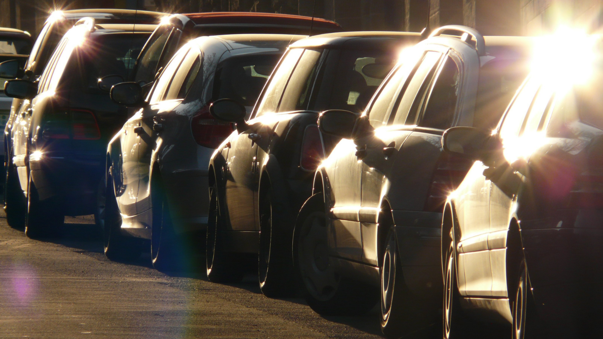 A line of cars parked nose to tail along a road, in the sunlight.