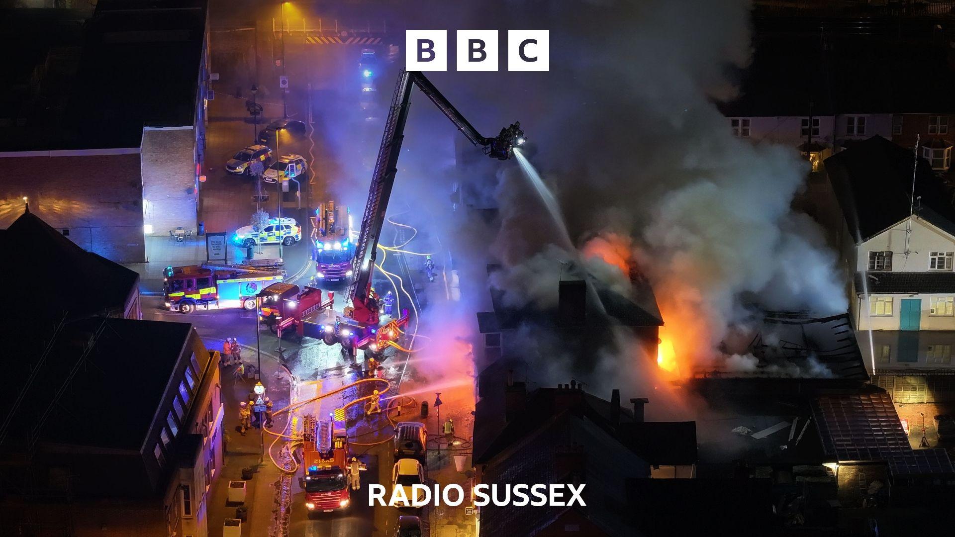 A night time aerial scene of fire engines tackling a fire in a building. One engine has a crane platform from which water is being sprayed down on the fire below. Thick black clouds of smoke fill the image.