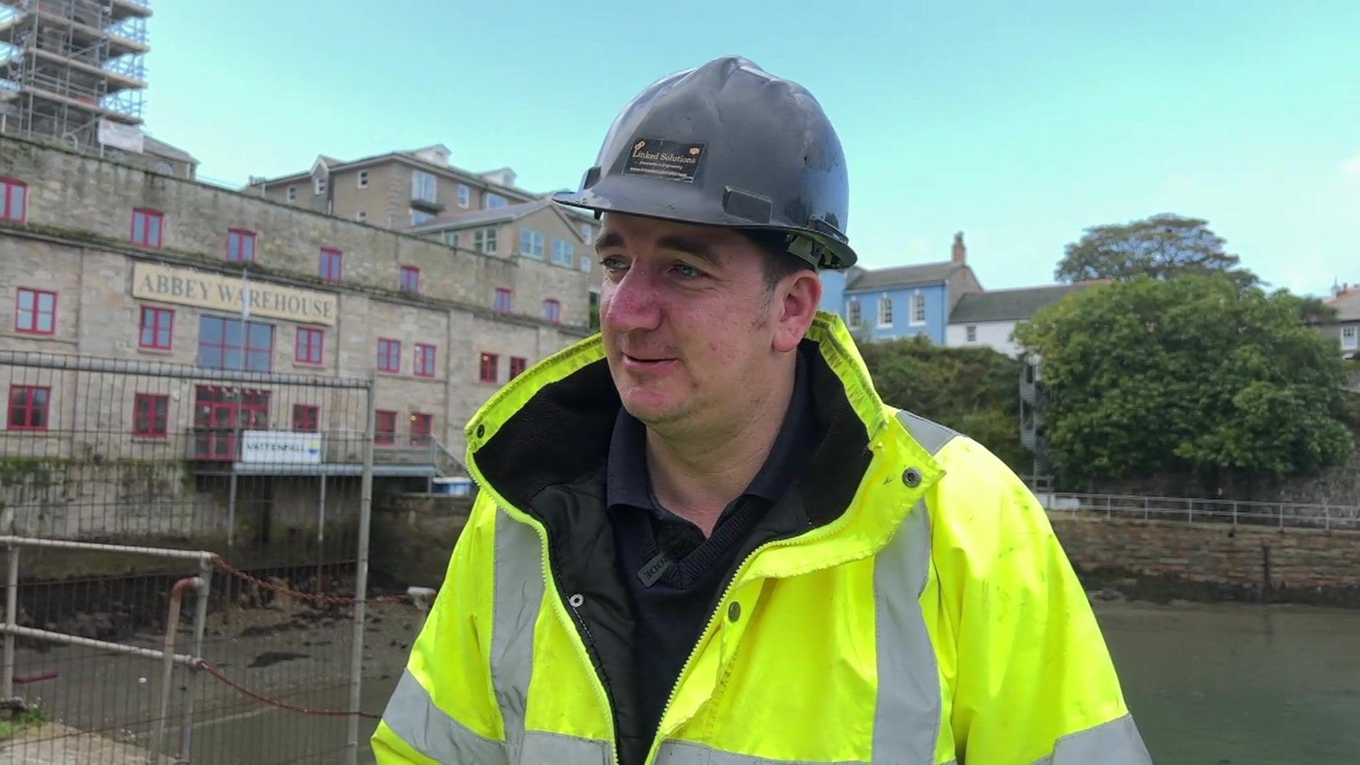 Jamie Murphy pictured wearing a yellow high visibility jacket with a navy blue jumper beneath. He is also wearing a dark grey hard hat and is clean shaven. He has been photographed in front of Abbey Warehouse. 