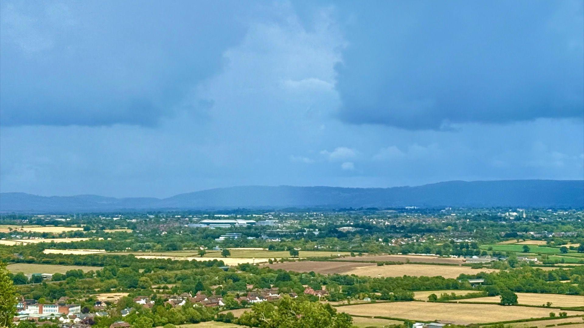 A view of fields and trees with a blue sky. In the background a range of hills, showing dark blue and grey, is visible