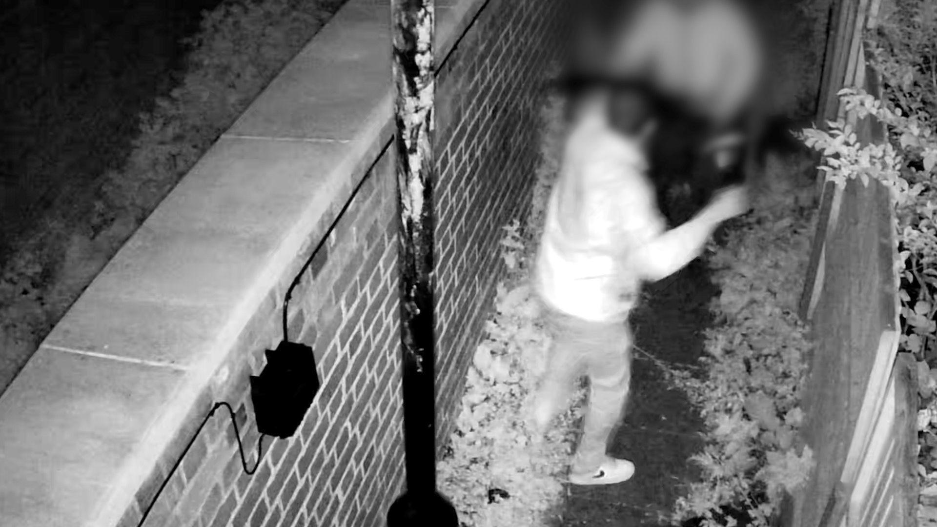 A black and white CCTV image shows a blurry image of a man in a jacket, jeans, and trainers holding up a hammer at another individual who is carrying a TV. They are both in an alley.