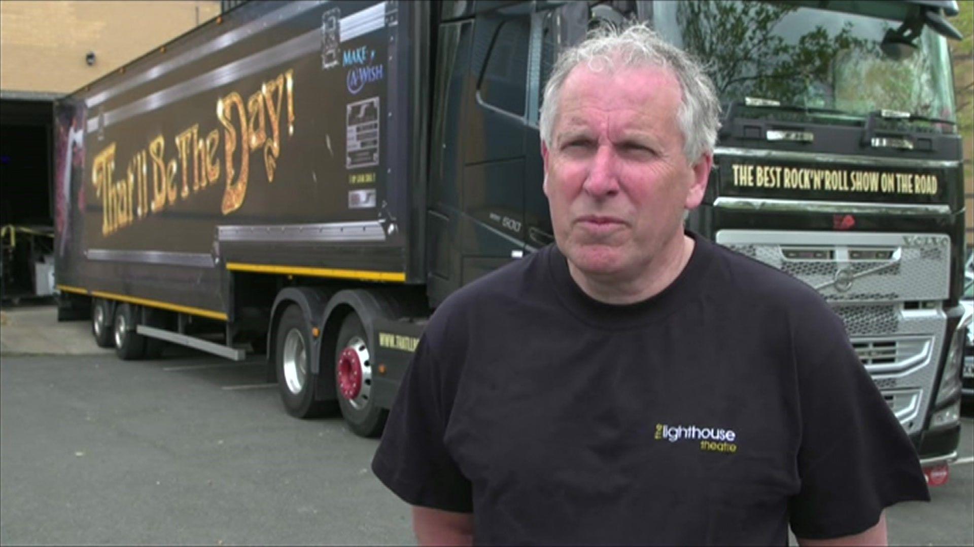 A man in a black T-shirt with "The Lighthouse Theatre" written on it, stands in front of a black lorry by the side of the theatre. He is unsmiling. 