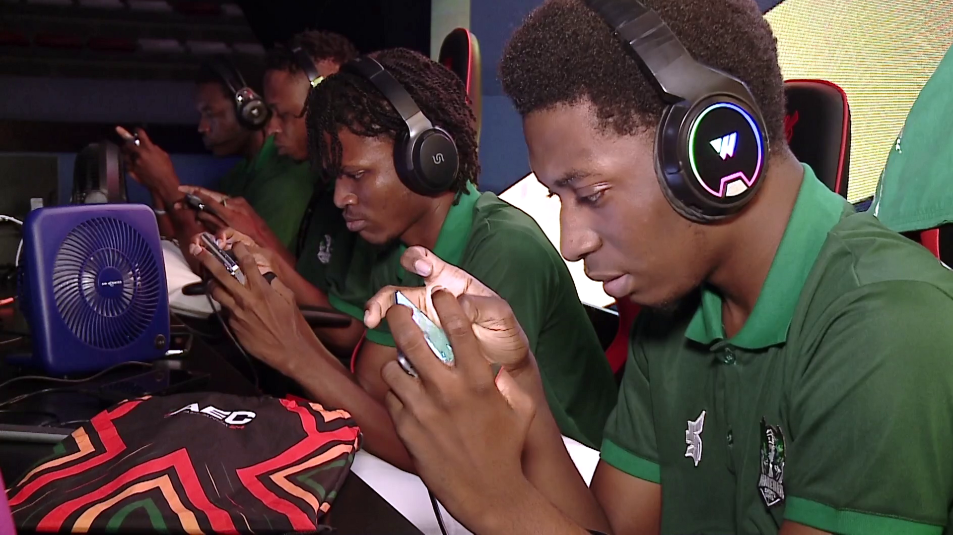 Several gamers, wearing headphones and matching shirts, concentrate on handheld consoles during an esports tournament