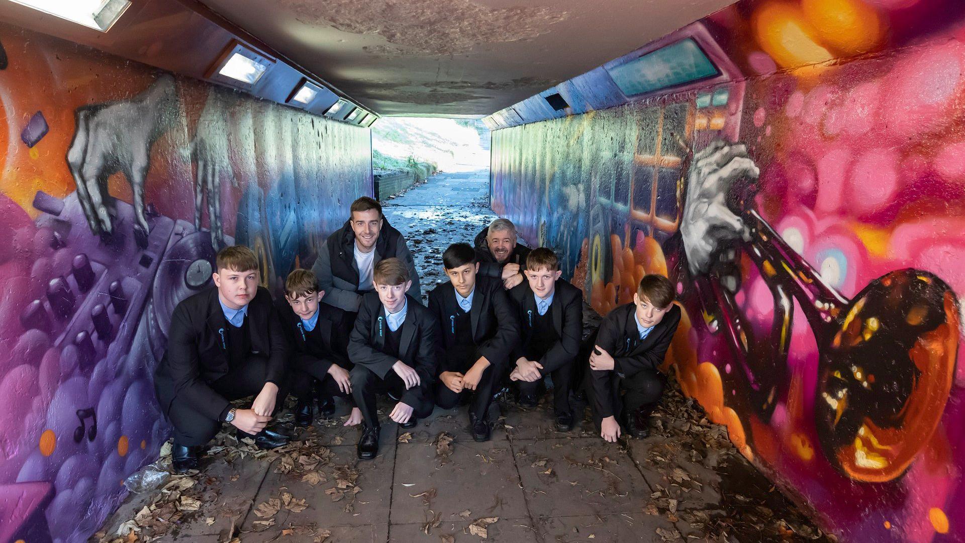 Five pupils from Studio West school, wearing their uniforms, in one of the underpasses along with artist Mark Shields and fellow artist Toby Heaps. The underpass is decorated with murals including a DJ mixer on the left-hand side of the image and a trumpet on the other.