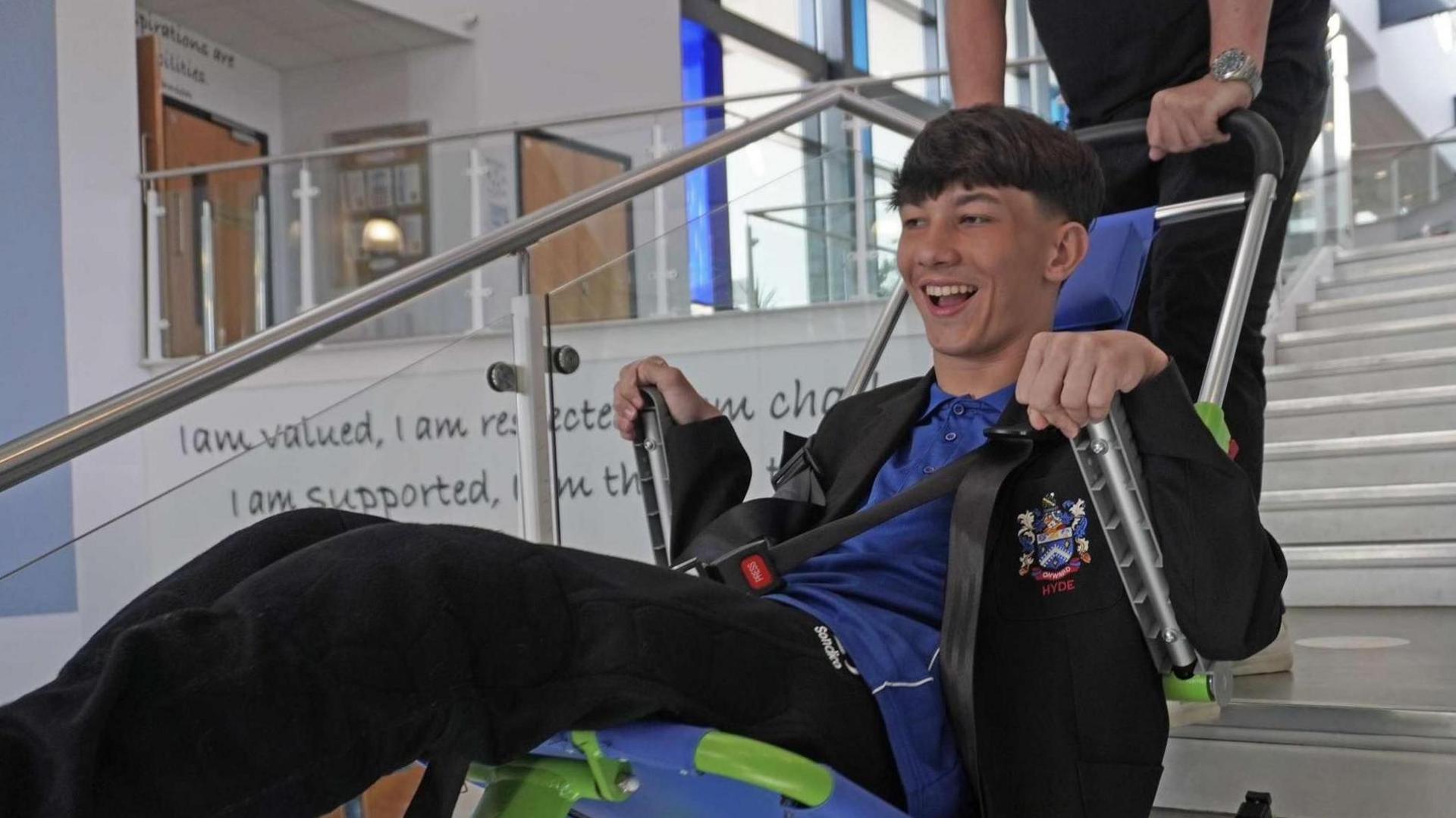 Lucas with brown hair wearing a school uniform smiles as he is lowered down the stairs of his school in an evacuation chair.