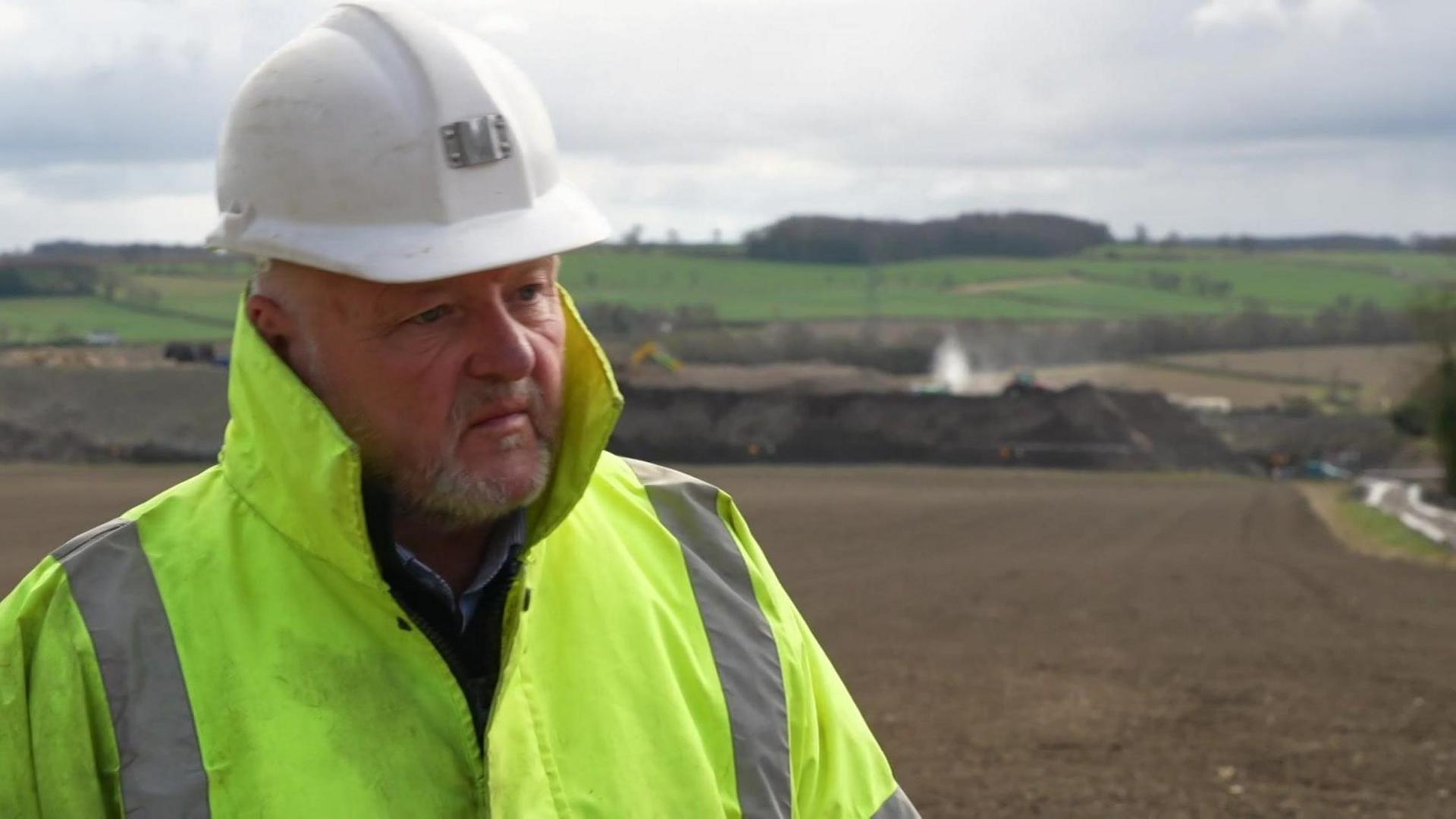 A man in a yellow hi-vis jacket and white hard hat stands on the quarry site 