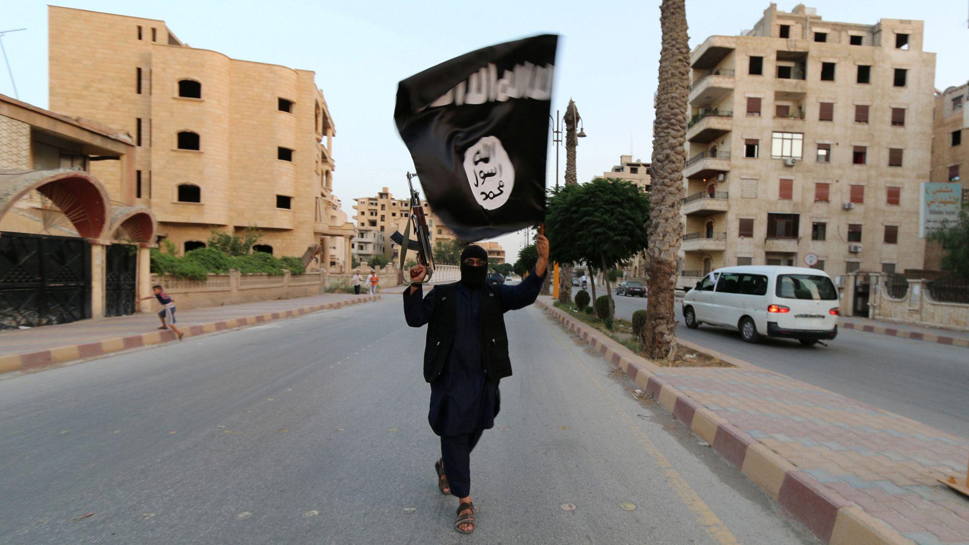 Member of IS in black clothing walking down a road in Raqqa holding a gun and IS flag, June 2104