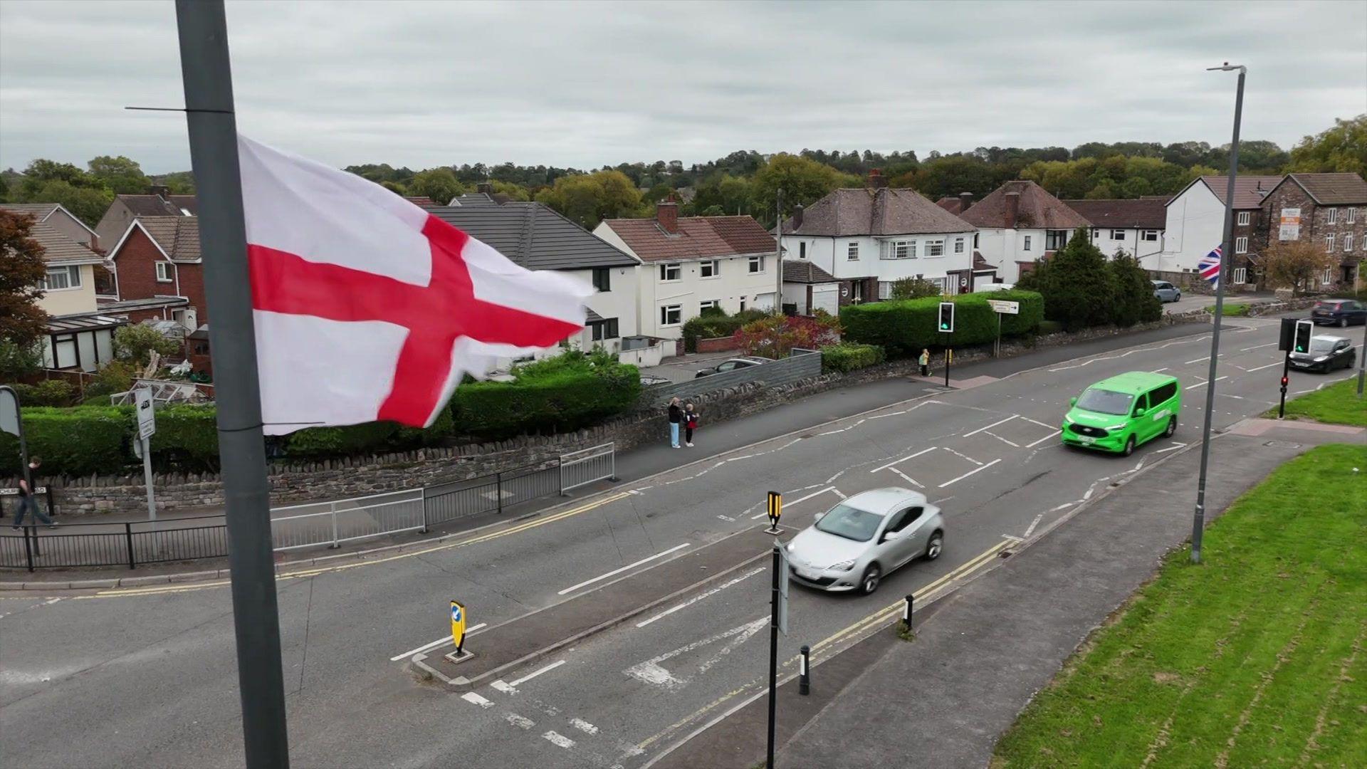 An England flag on a lamppost in South Gloucestershire. Houses can be seen in the distance and there are cars driving along the road.