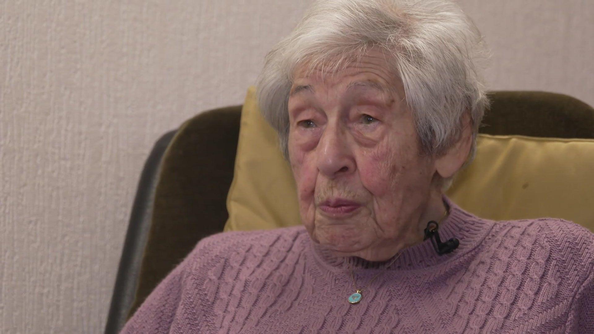 An elderly woman wears a pink knit jumper and looks to the left of the camera as she sits in an armchair