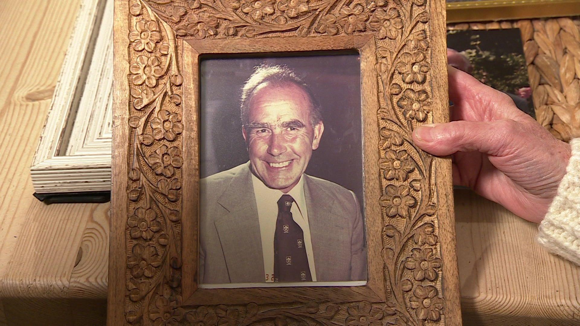 A person holds a framed photo of a man. The man is smiling at the camera. He has short grey hair and wears a grey shirt with a tie.