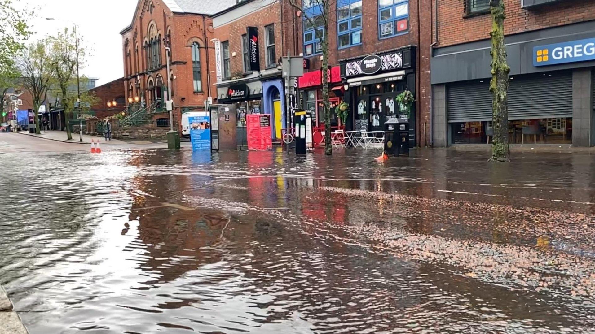 Flooding on Botanic Avenue. A large puddle of water is on the street with shopfronts in the background.
