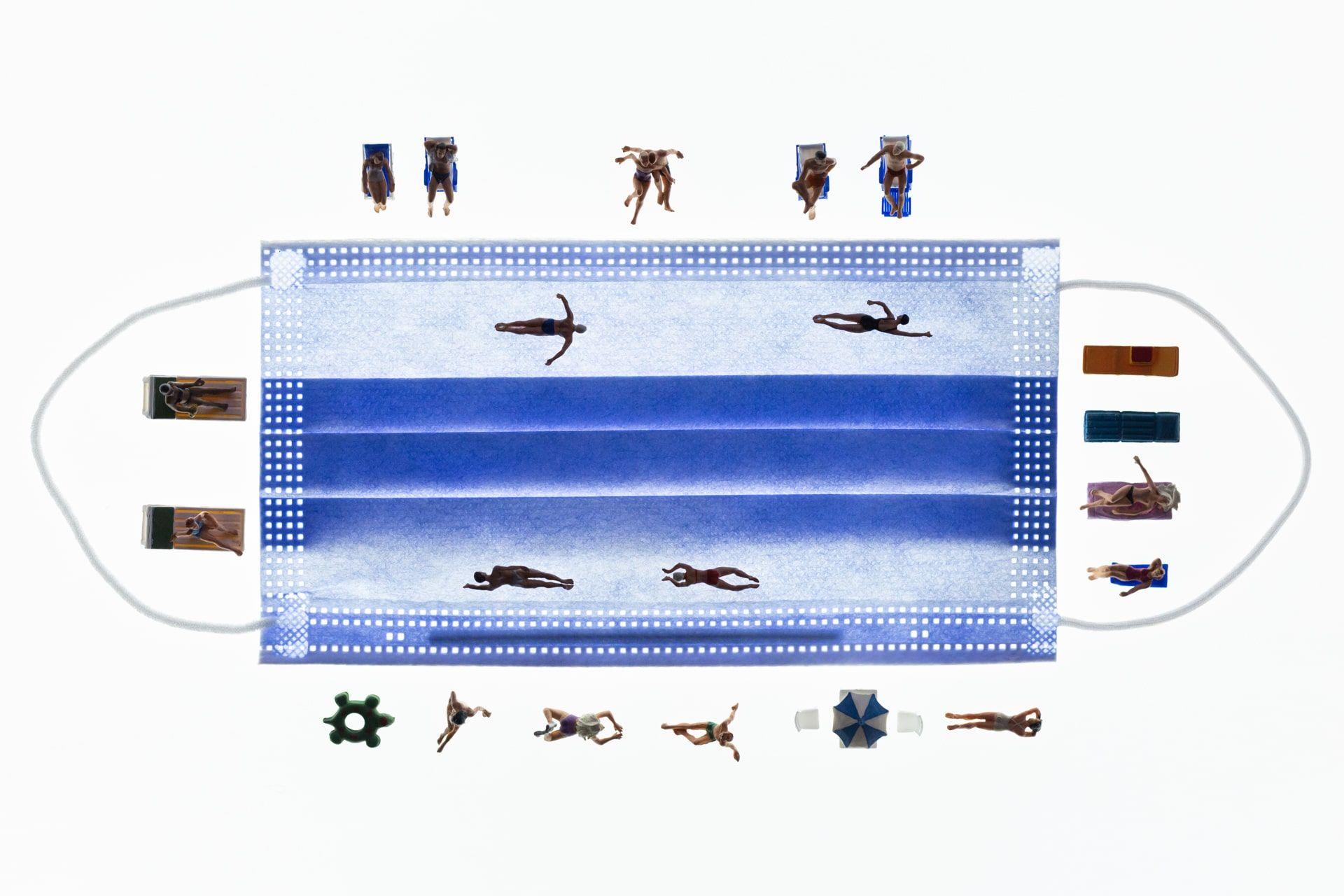 A blue surgical mask is against a white background, which looks like lands of a swimming pool. There are small figurines sunbathing around it along with others that look like they are swimming in the mask. 