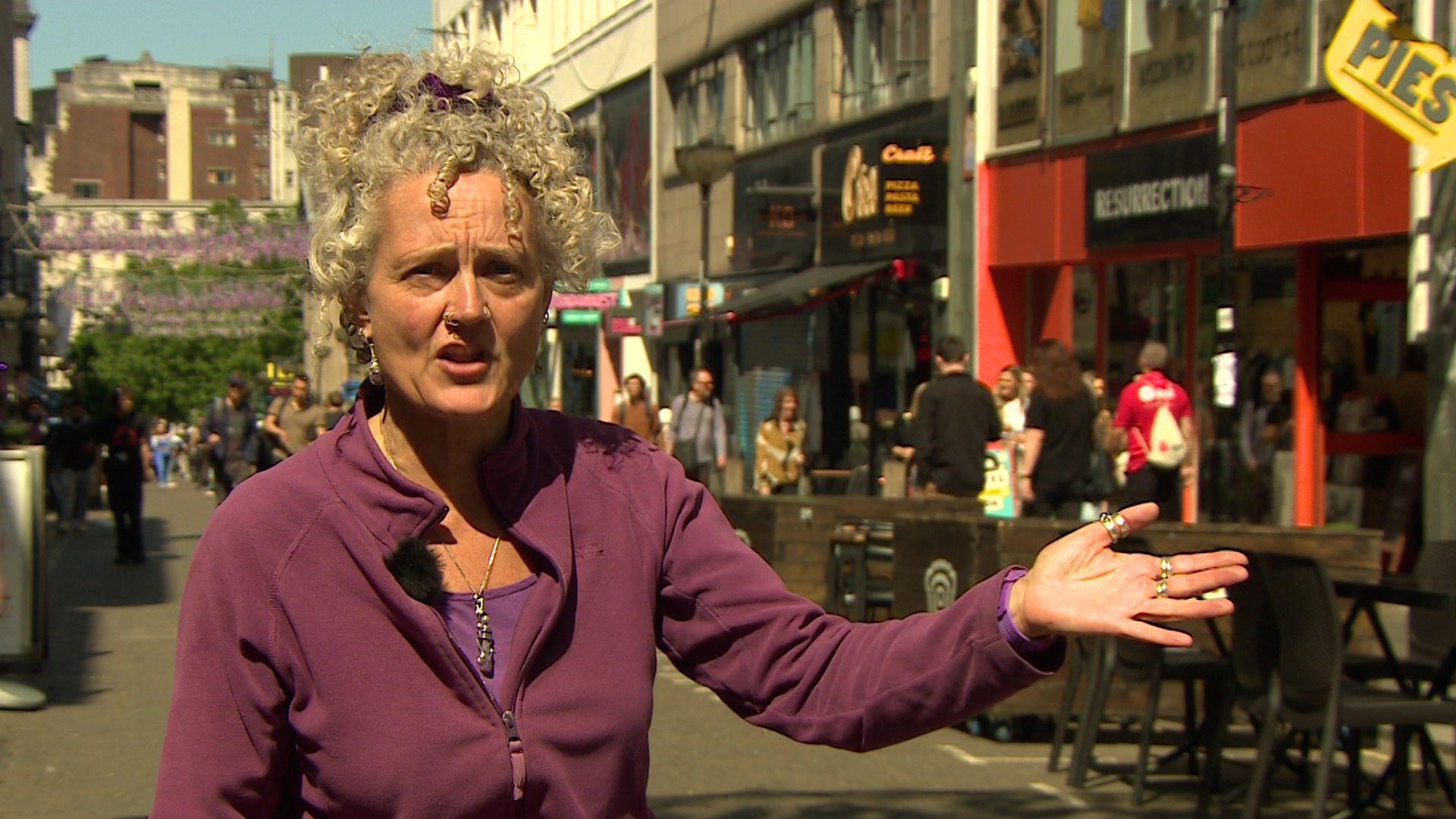 Dr Kay Hinkle, wearing a purple fleece, speaks to the camera against a backdrop of shops and restaurants in Bold Street