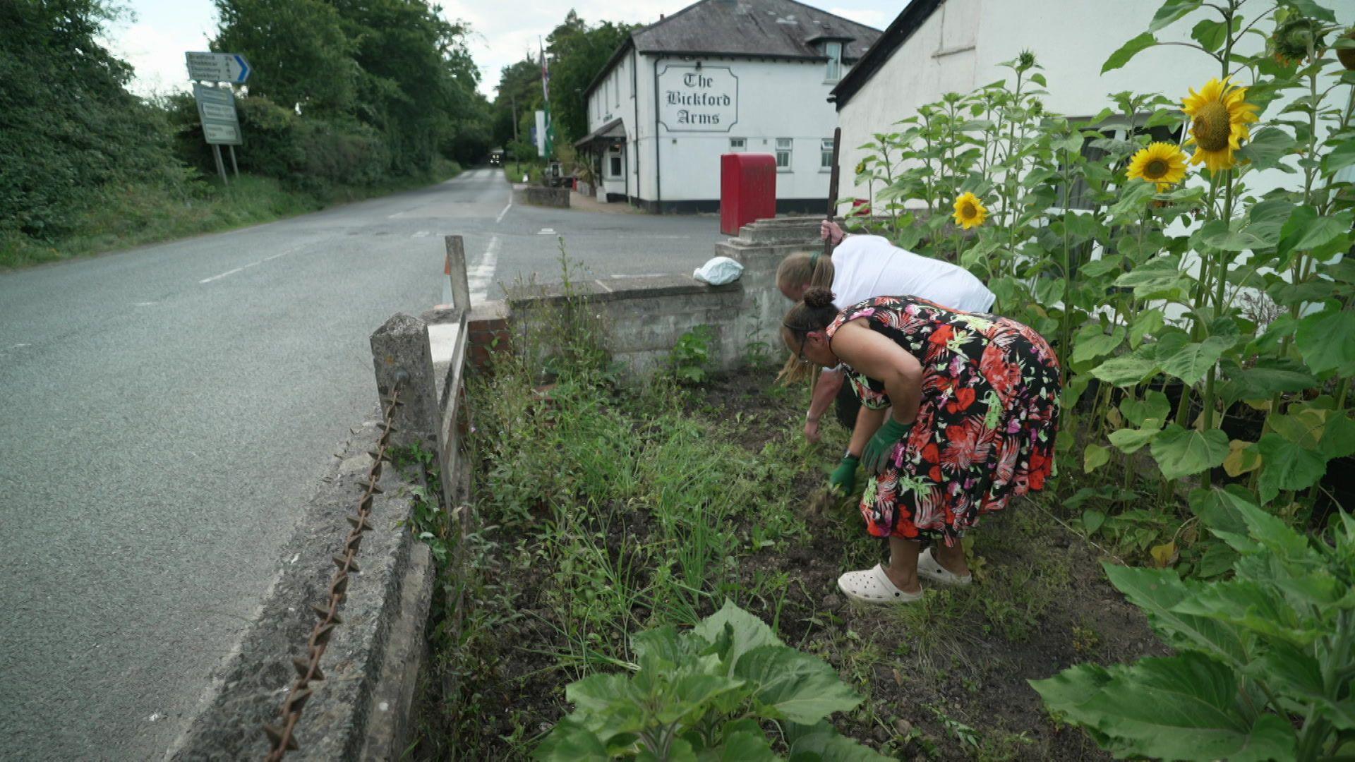 Shanae Boorman-Leech gardening with her husband in their garden. Its walls are on the road and in the background is the local pub, The Bickford Arms.