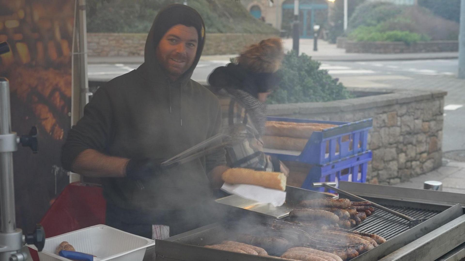 A man wearing a hoodie and food-preparation gloves is grilling sausages on an outside grill. One hand is holding a pair of tongs and the other half a baguette. The smoke from the grill rises all around him.