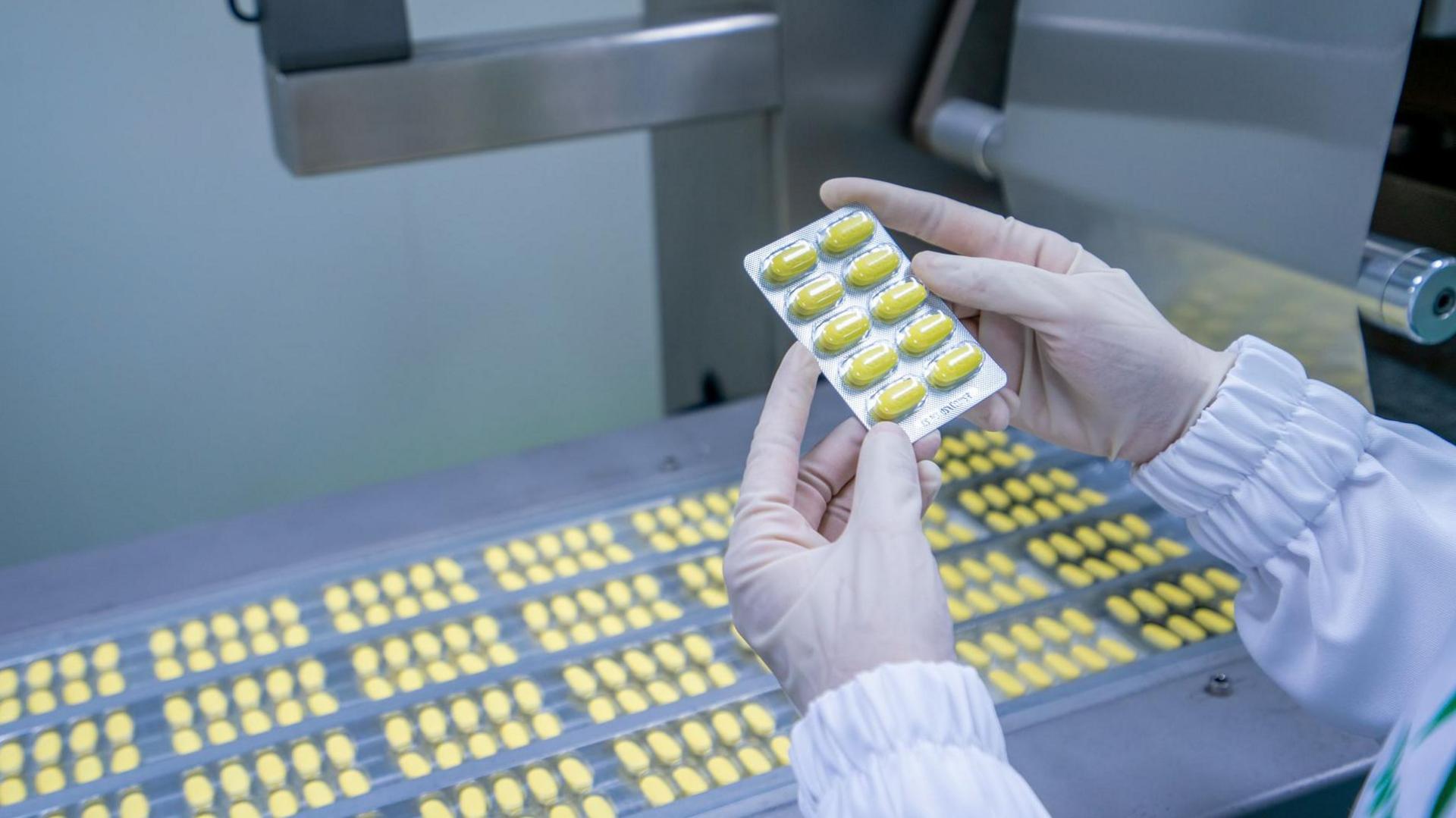 Gloved hands hold a tray of yellow tablets, above rows of yellow tablets on a conveyor belt.