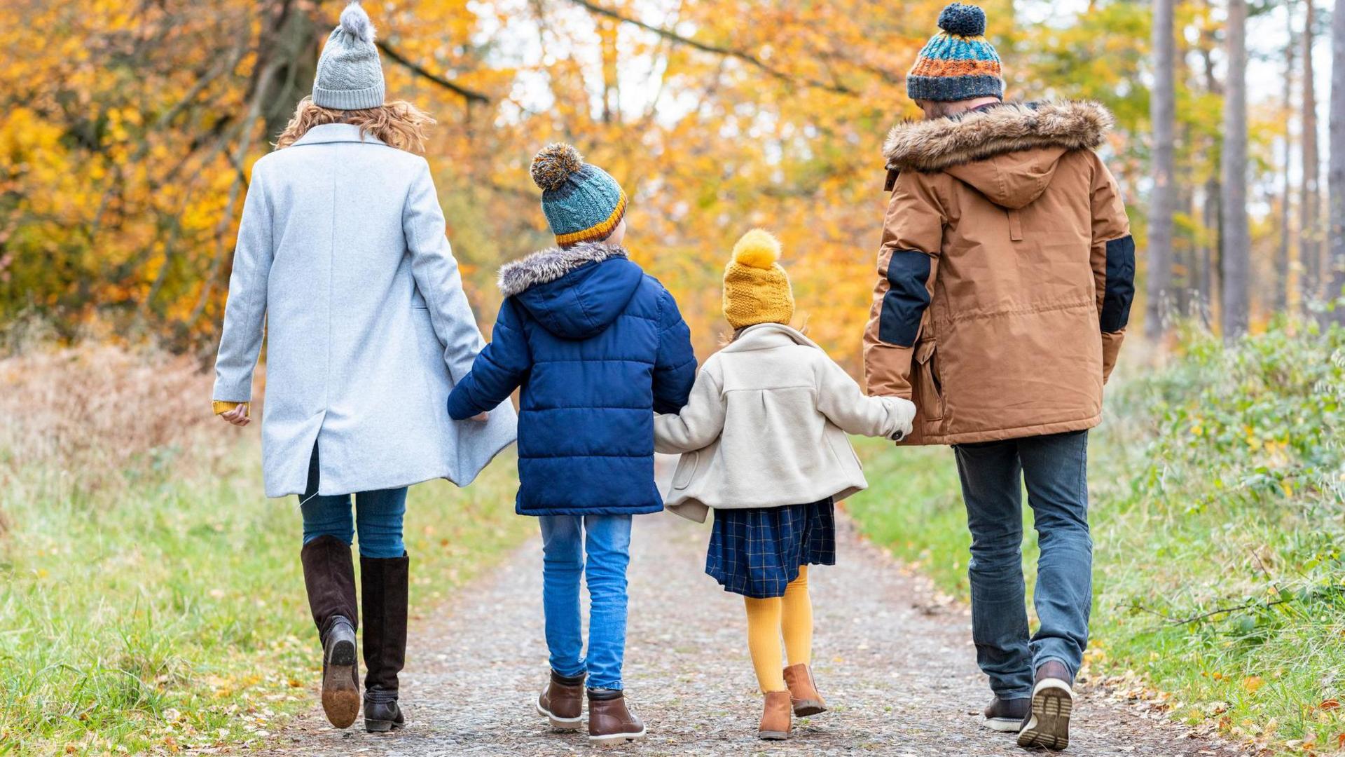 A stock image of a family walking on a woodland path. On the right a woman wearing a winter coat, a bobbly hat and knee-high boots. Next, a boy wearing a blue park, a bobbly heat and jeans. He is holding hands with a young girl who is wearing a yellow hat and tights, with a coat and blue dress. On the right is a man wearing a hat and a brown parka jacket.