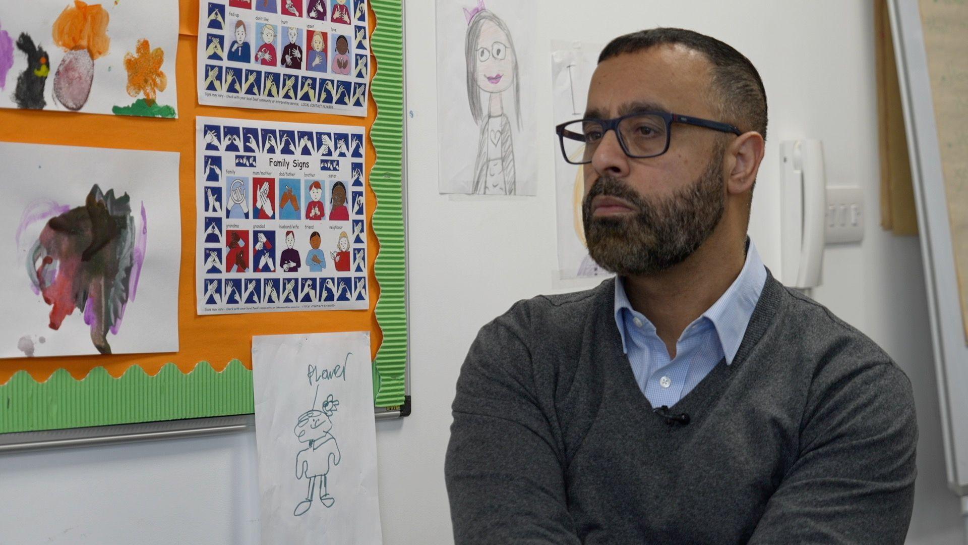 A man with glasses and a short beard is looking straight-faced in the left of the camera. His blue shirt collar is visible above his dark grey jumper. He is sat to the right of the picture next to a display board of children's art and British Sign Language outlines