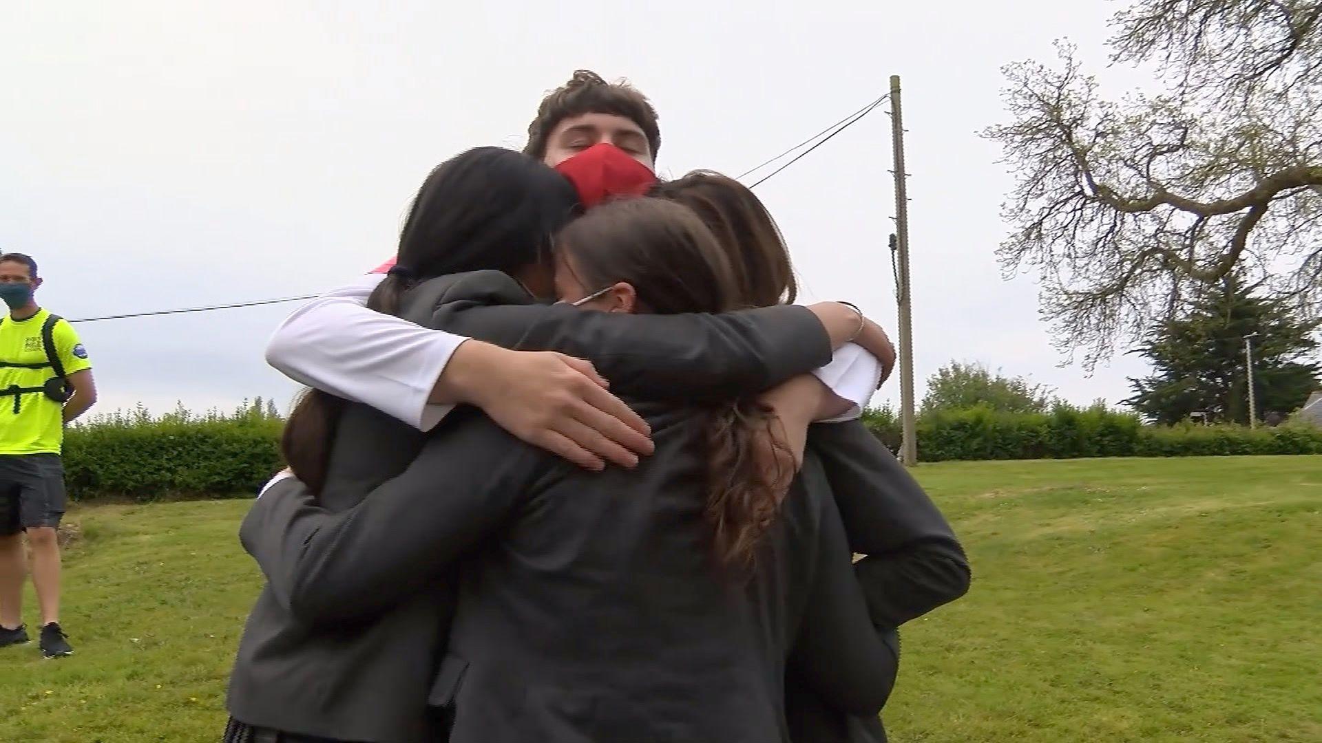 Four children in grey school blazers with their arms around each other in a group hug, in a grassy field. Fintan can be seen taller than the others, wearing a red face mask.