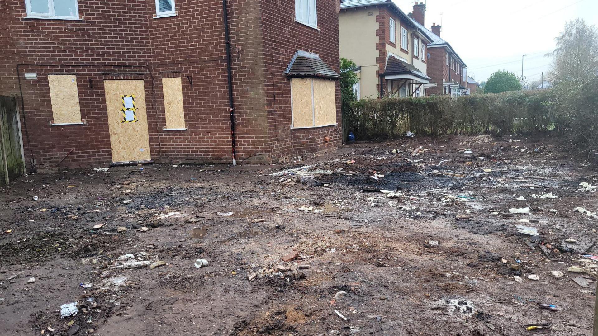 A house, with windows and doors boarded up, can be seen with a large patch of mud in front of it. There is a paper notice taped onto one of the boards with yellow and black tape. There are neighbouring houses visible in the background, behind a privet hedge.