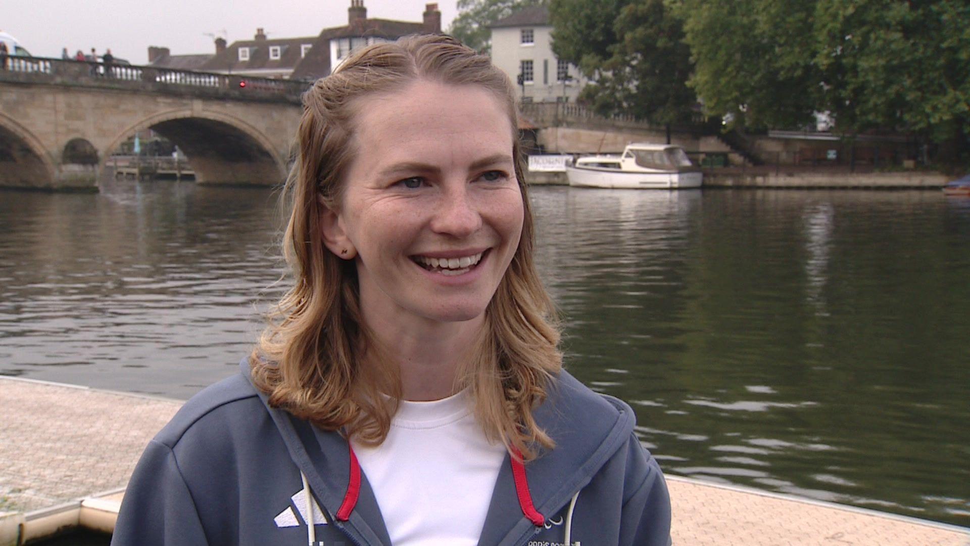 Erin Kennedy is dressed in a grey, zip-up Adidas hooded top and is wearing a white T-shirt underneath that. She has shoulder-length mousey brown hair and is smiling. She is stood in front of the River Thames in Henley, with a bridge and a boat behind her 