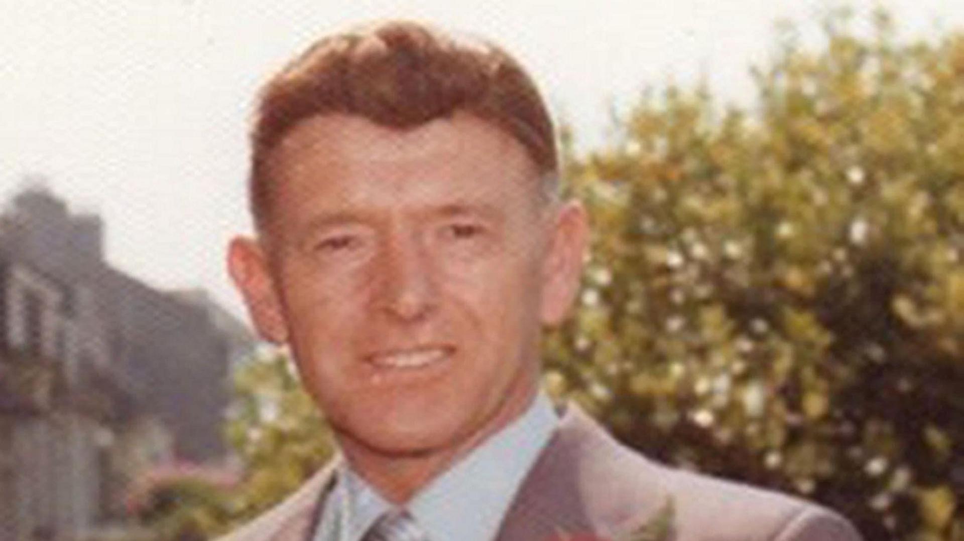 George Murdoch looking directly at the camera. He has dark eyes and brown hair and is wearing a grey suit with a light blue shirt.