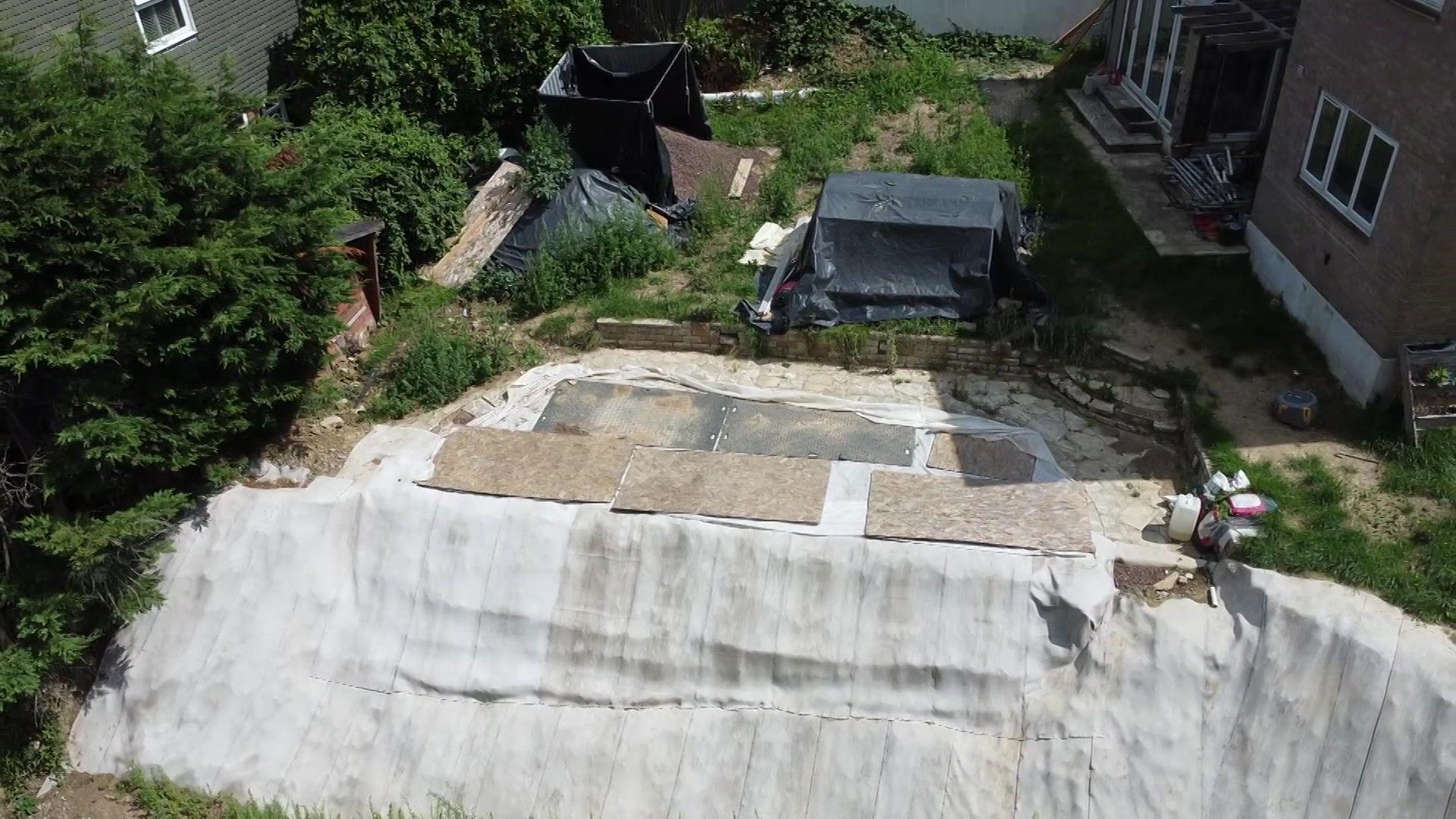 A white tarpaulin covers part of a garden, with trees to the left and a house to the right