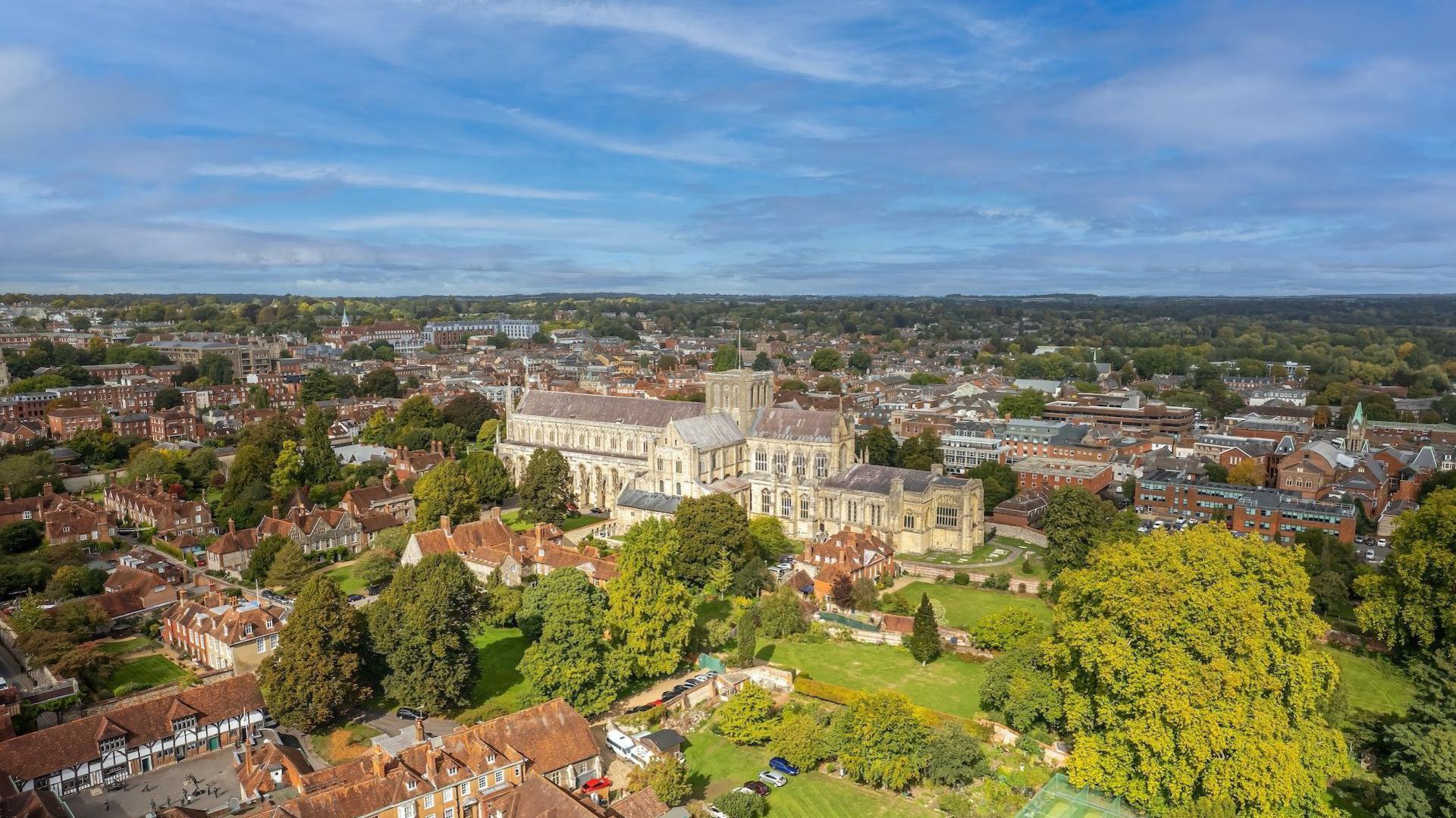 An aerial view of Winchester. The large stone cathedral building is surrounded by homes. In the foreground there is a large tree park area with trees.