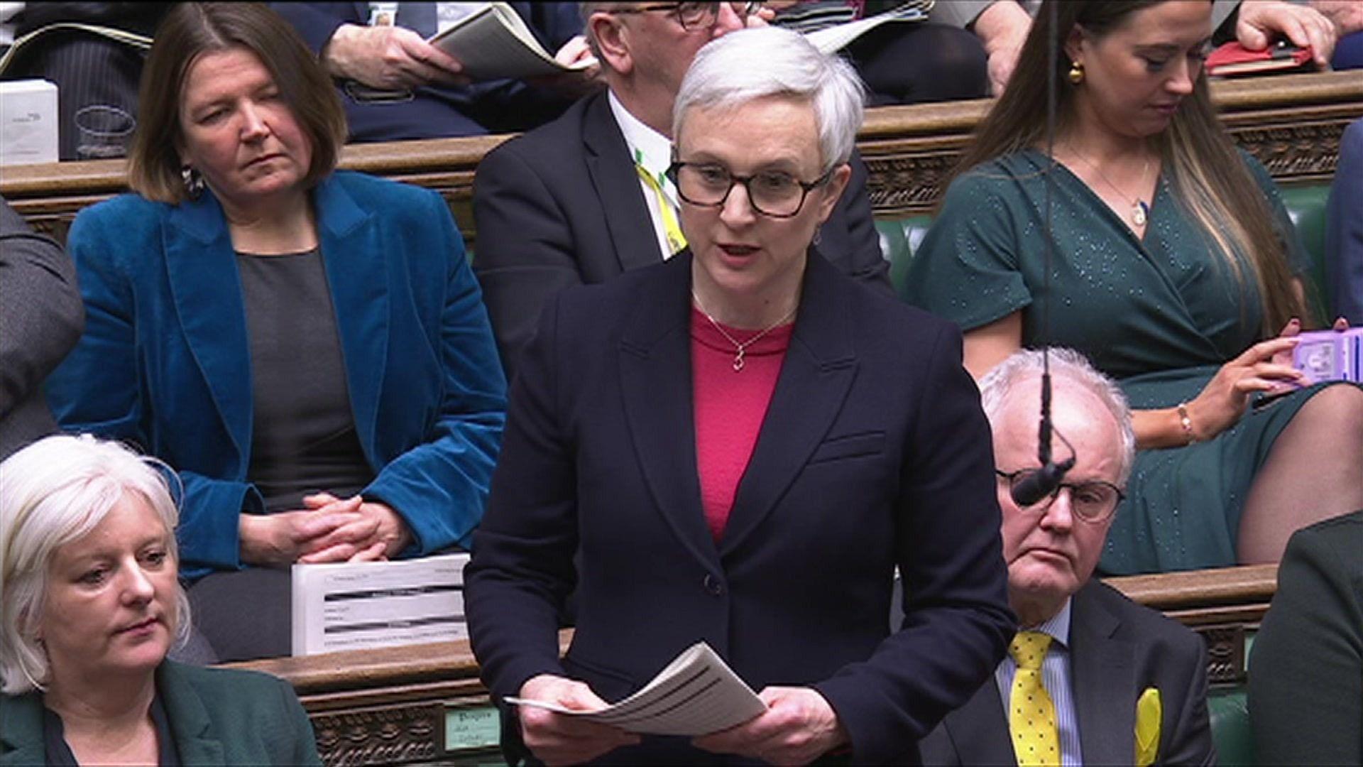 Alex Brewer MP is standing up in the House of Commons to ask the question. She has short grey hair brushed to the side, dark-rimmed glasses, and is wearing a black blazer with a red top underneath. She is holding some documents and is surrounded by her MP colleagues.