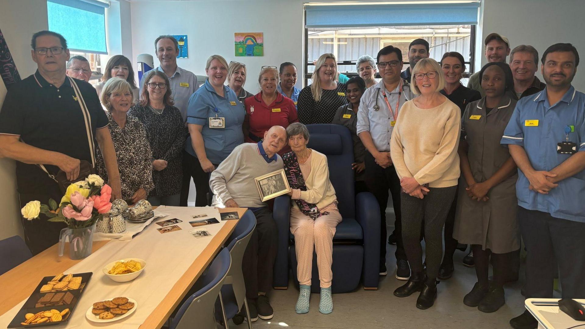 A large group of people including family members and hospital staff surround Elizabeth and John Ross and smile at the camera. 