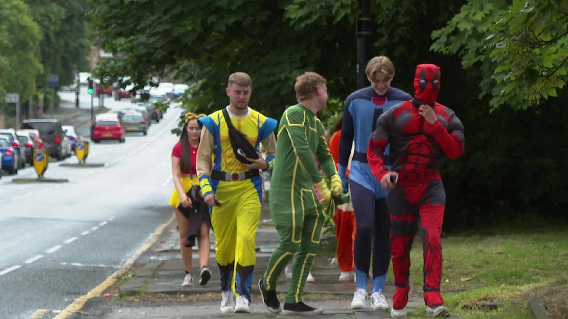 A group of young people in superhero fancy dress outfits walk down a city street.