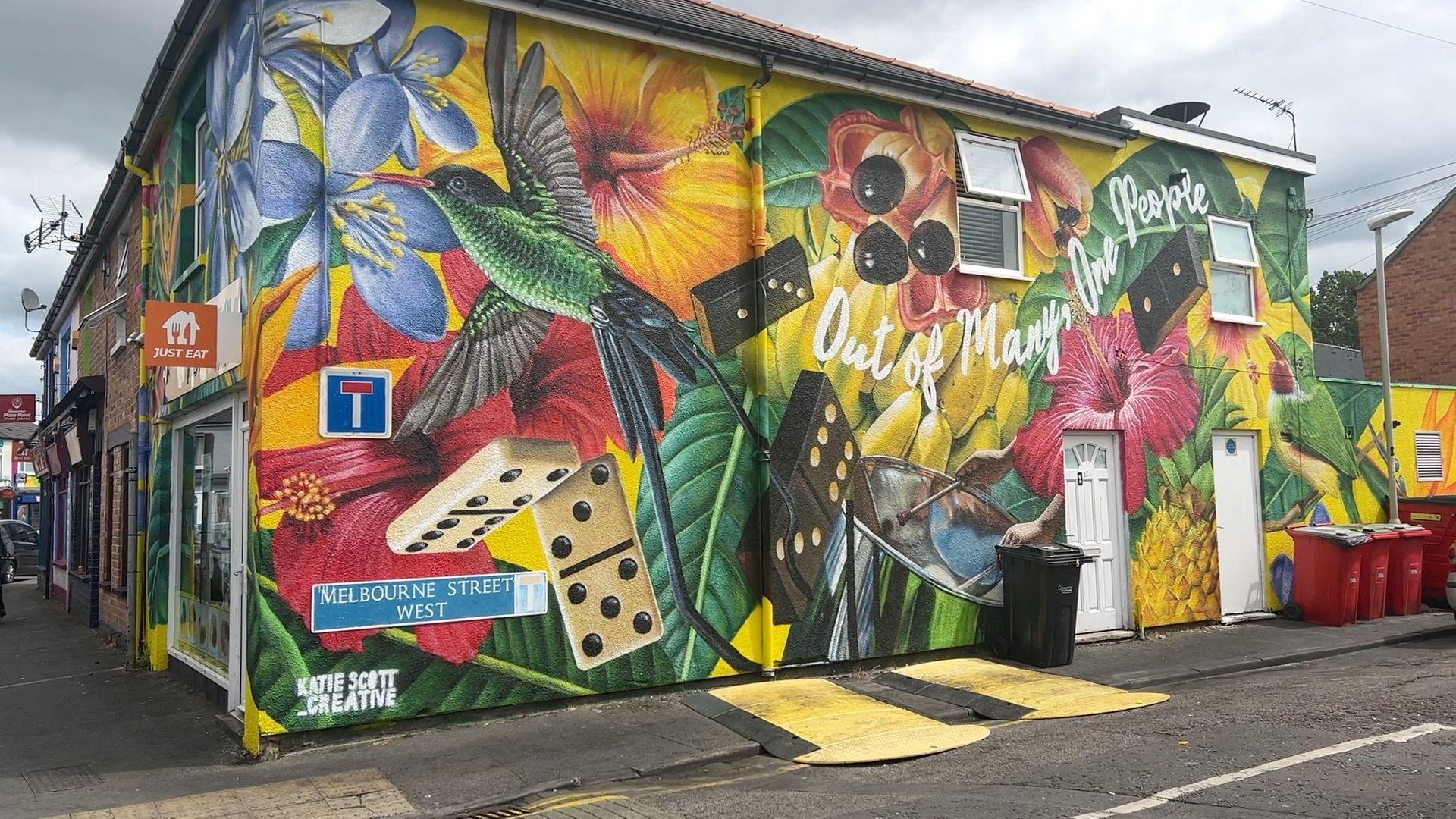 Mural painted on a building. It is colourful, and features dominoes, flowers, fruit and birds. A street sign for Melbourne Street West can be seen on the bottom left hand corner of the building. 