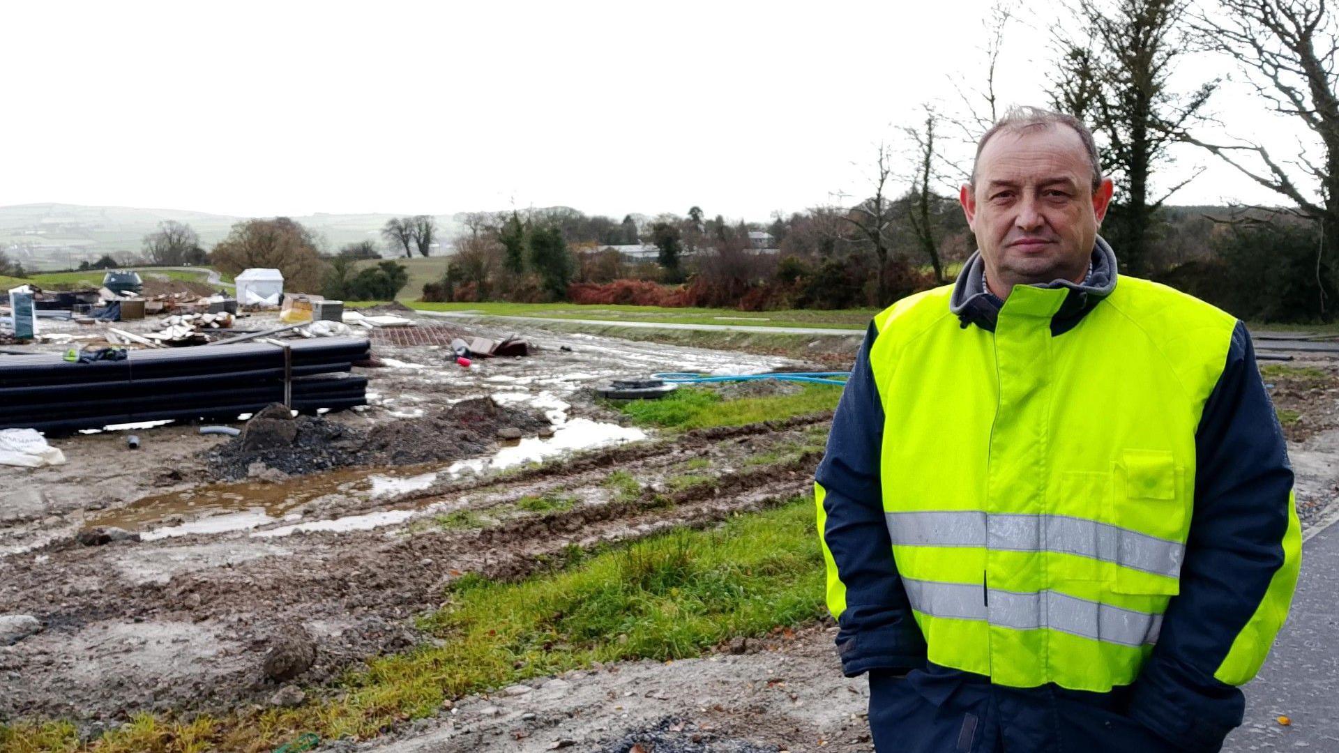 Councillor Declan Norris stands with his hands the pockets of a hi-vis vest cut off at the shoulders, showing the dark sleeves of the fleece he wears underneath. Behind him is what looks like waste ground, where the imprint of tire tracks from heavy machinery can be seen.