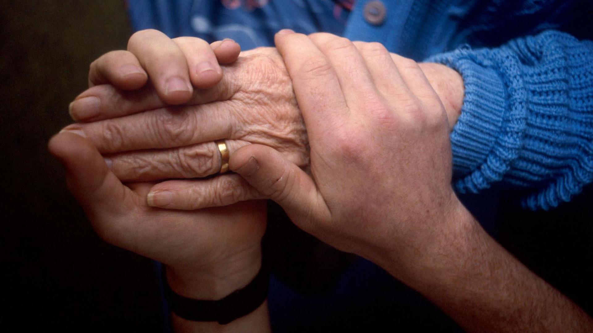 A younger person holds the left hand of an elderly person who is wearing a blue knitted cardigan and has a gold wedding ring on.