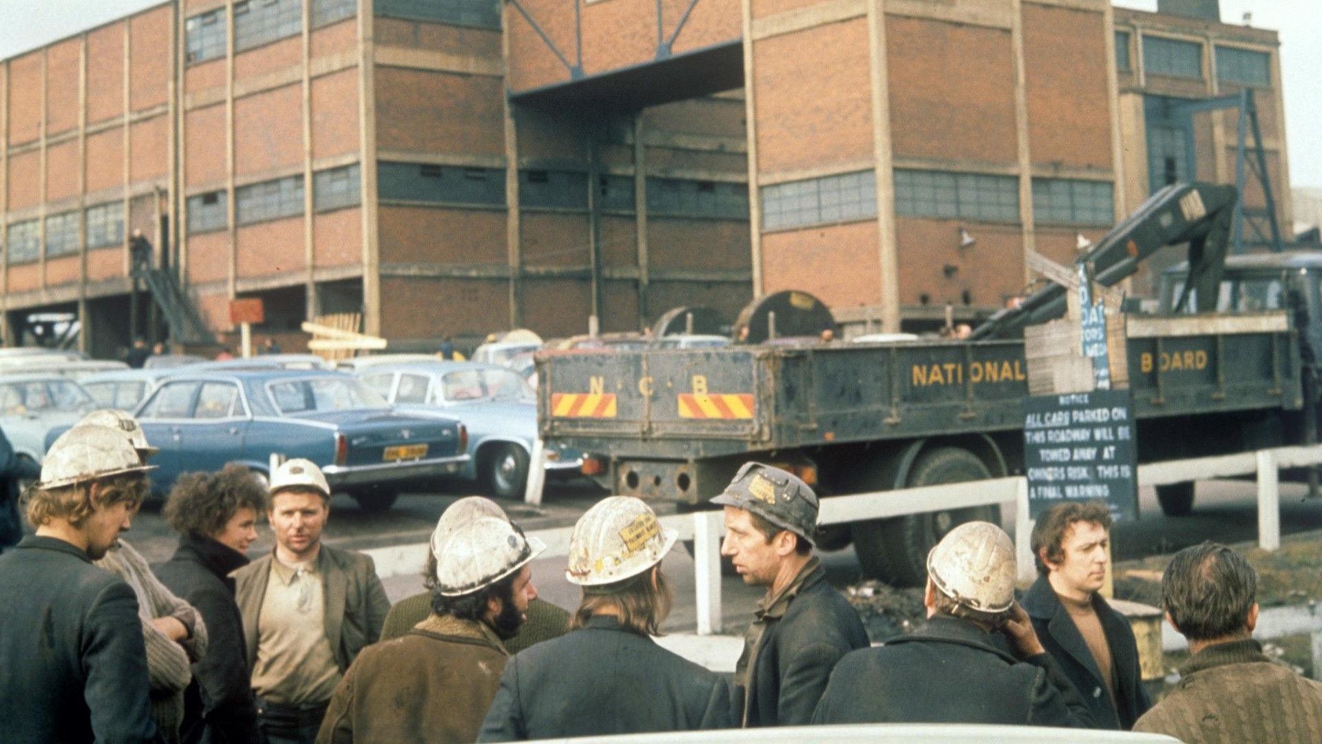 Archive image of miners wearing hard hats standing in front of a car park, with Lofthouse Colliery in the background.