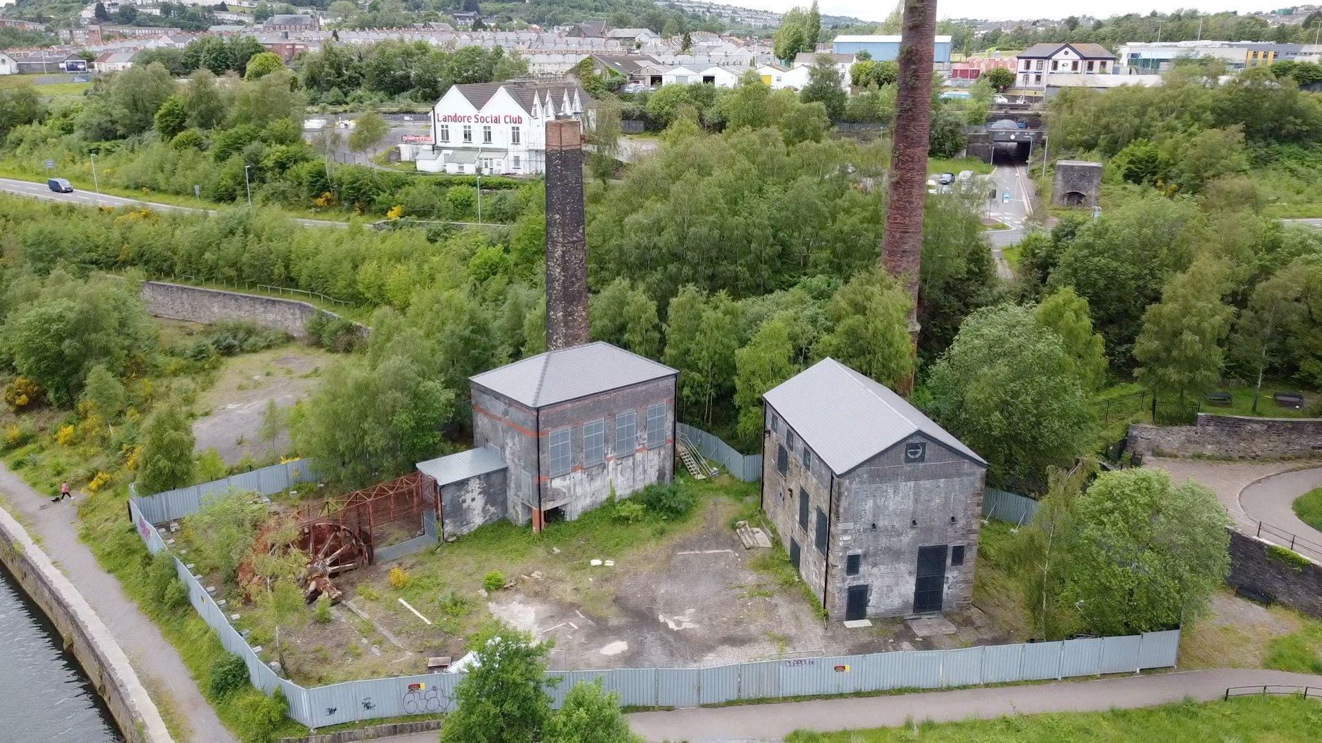 The engine houses looking in need of some restoration, with metal fencing surrounding them, as well as some rusty industrial equipment. Trees and shrubs can also be seen.