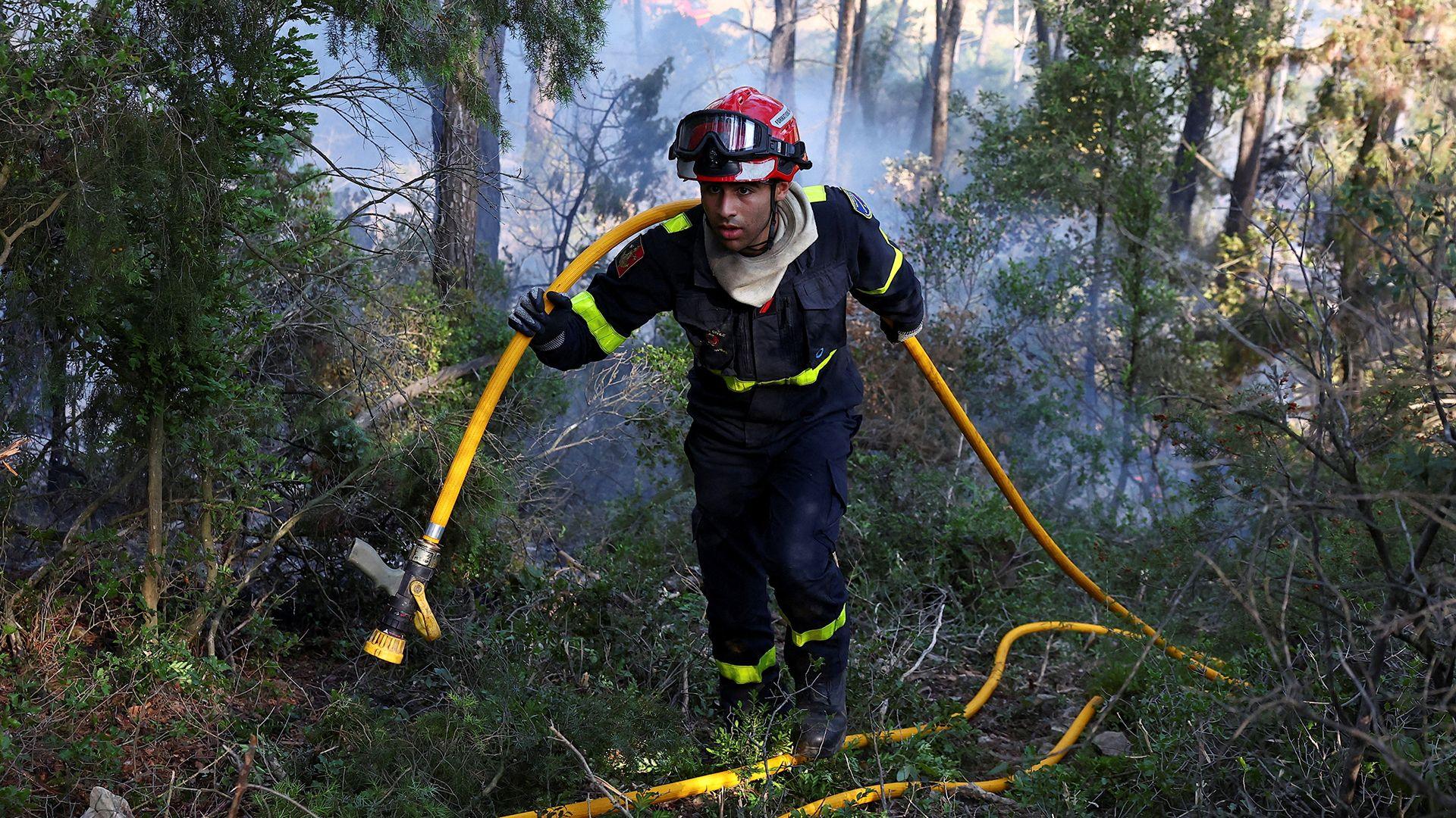 A firefighter holds a yellow hose over his shoulder as he walks through a burning forest area clouded with smoke.