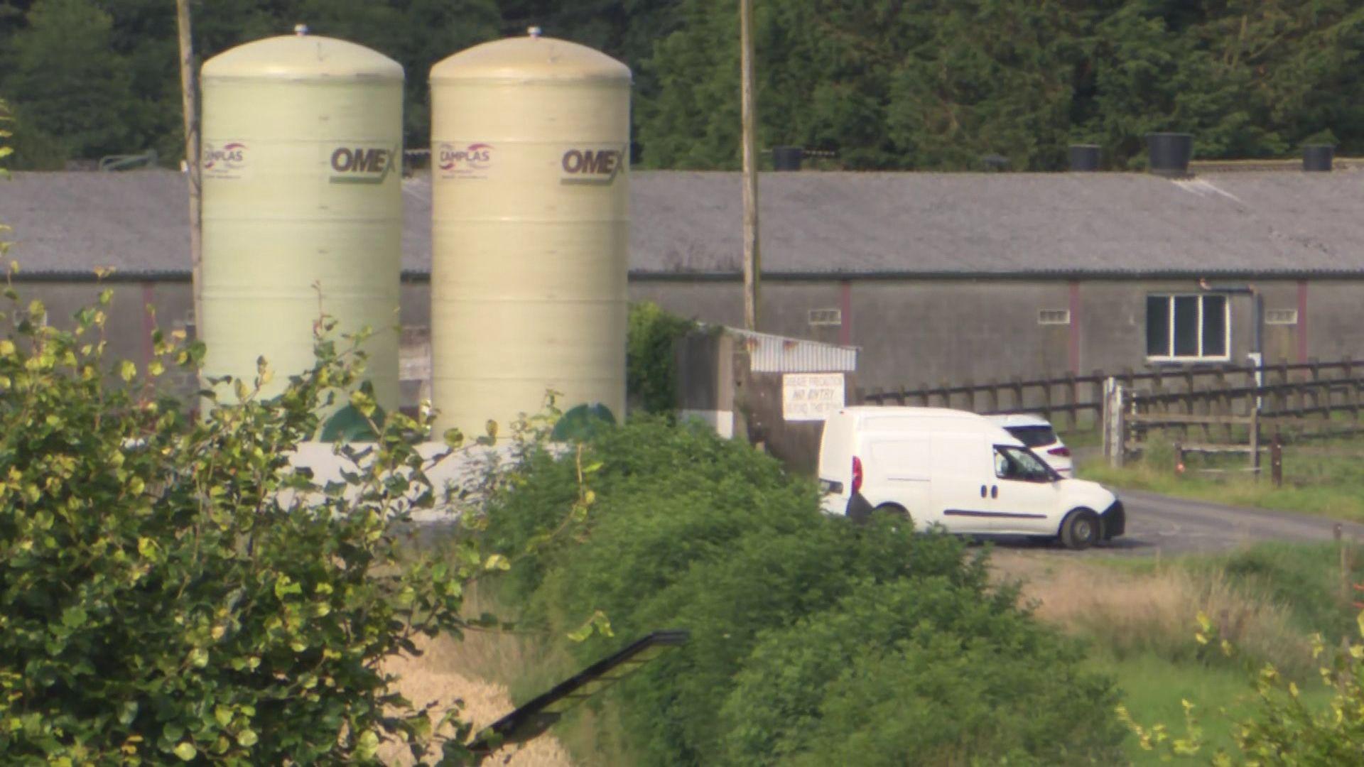 A white van and car parked by a brown fenced entrance to a grey building surrounded by green bushes and grass with large green and yellow tanks 