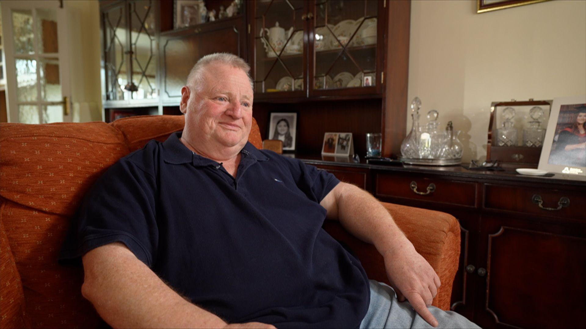 Ray, sitting in a red armchair in his living room wearing a black polo shirt, with family photos, fine china and a decanter set on the furniture behind him