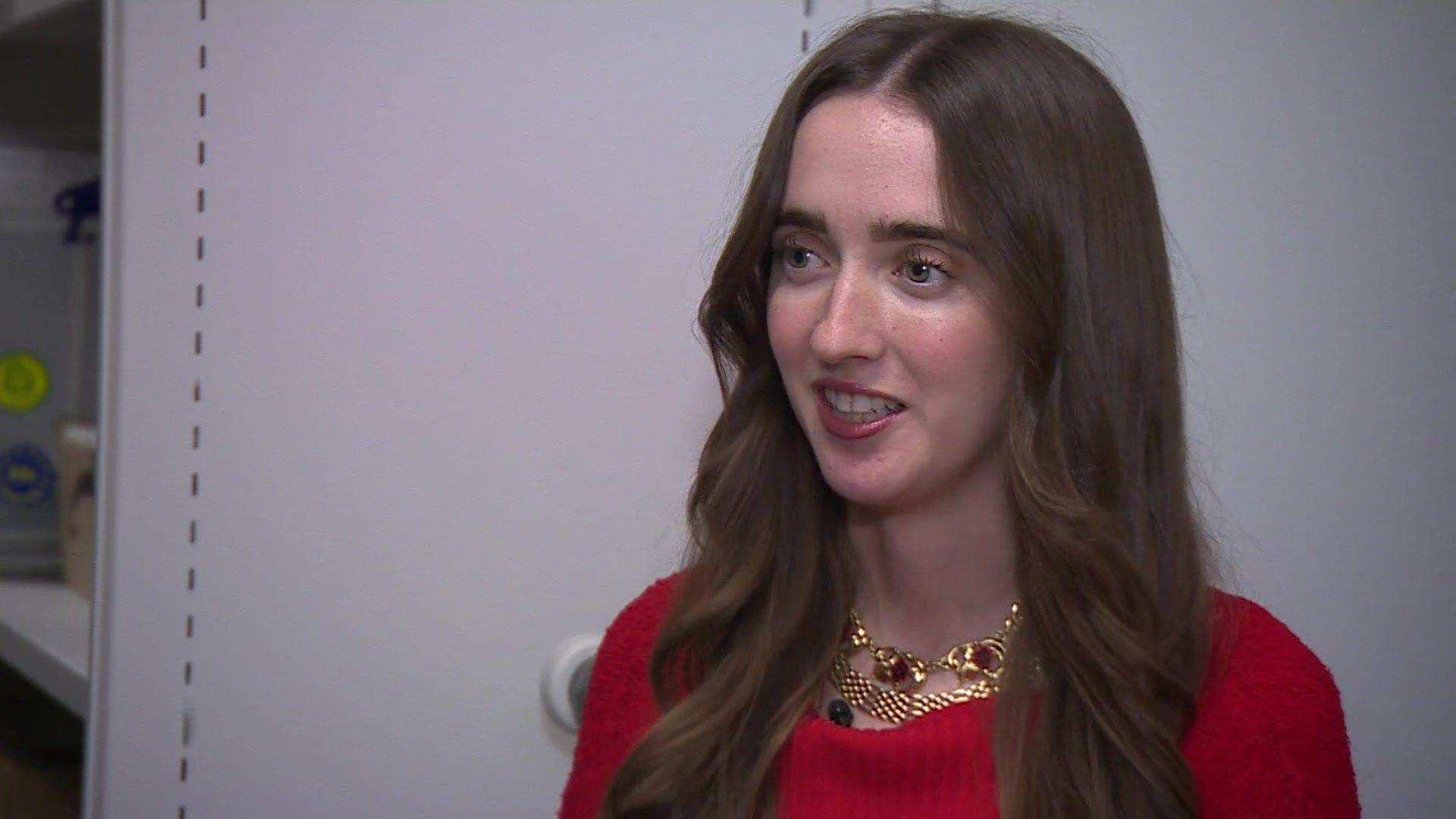 Holly Scott, wearing a red top, stands in front of some plain grey shelving used for storing theatre archives. The has long dark hair and wears a gold necklace with red stones.