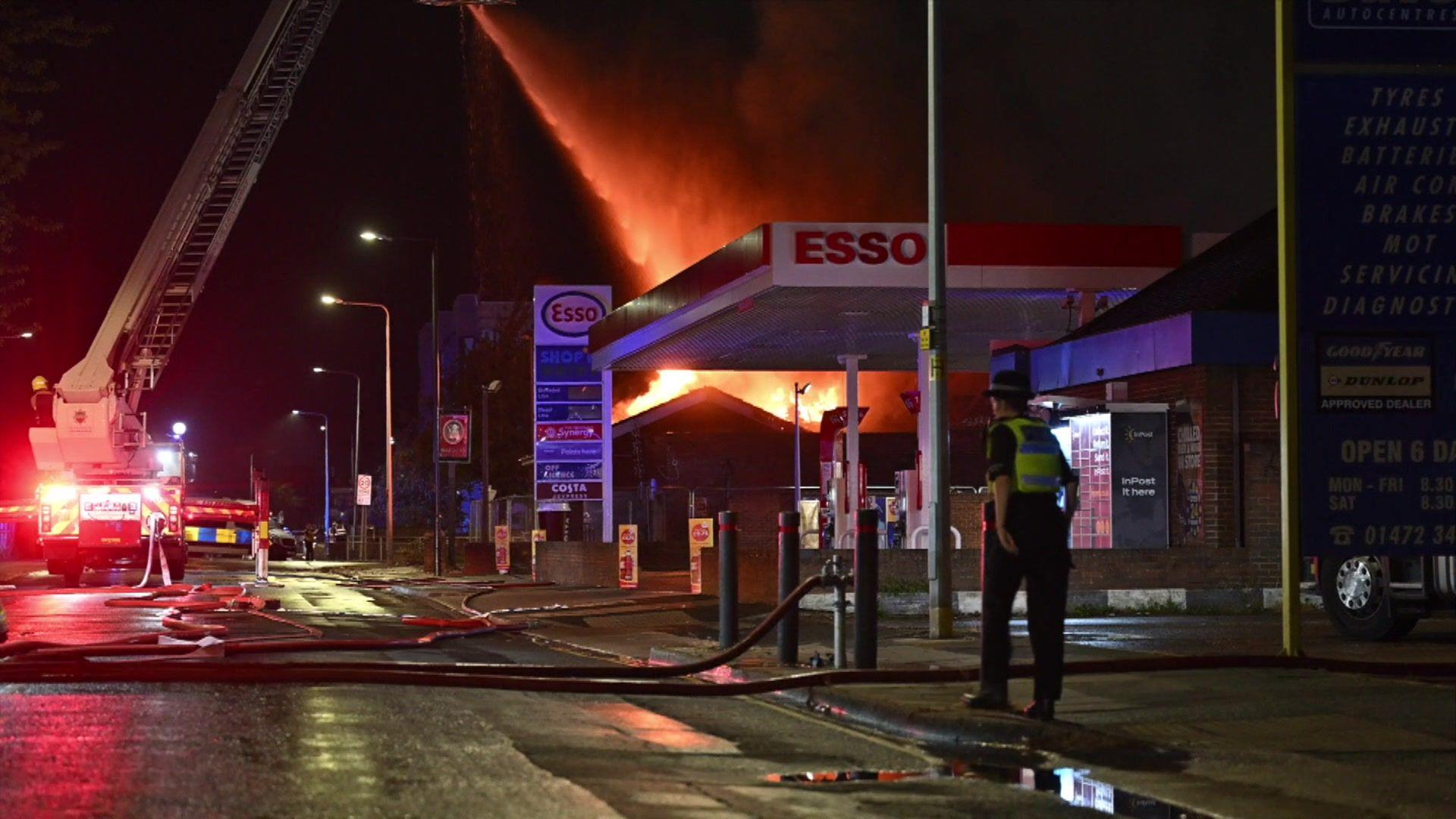 Water is sprayed from an aerial platform on to the roof of the former Mariners Rest pub in Grimsby. The night sky is glowing red and orange from a fire and a police officer can be seen in the foreground, close to a petrol station forecourt