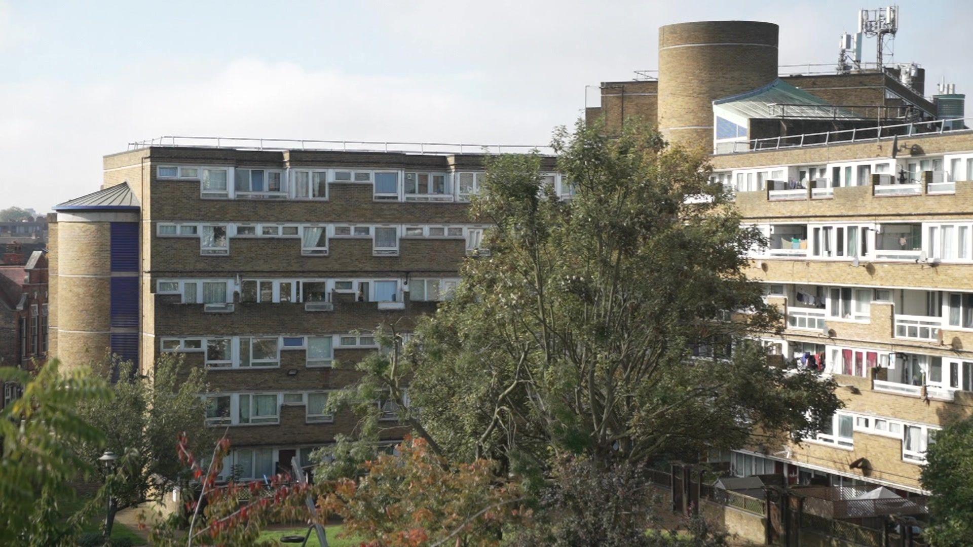 An image of the Gloucester Grove Estate in Peckham - comprising brick flats with communal walkways and a green space with trees near the flats