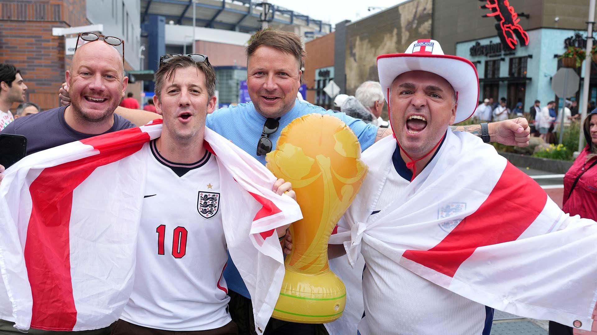 Four male England football fans draped in England flags outside Boston Football stadium. They have an inflatable world cup trophy with them.