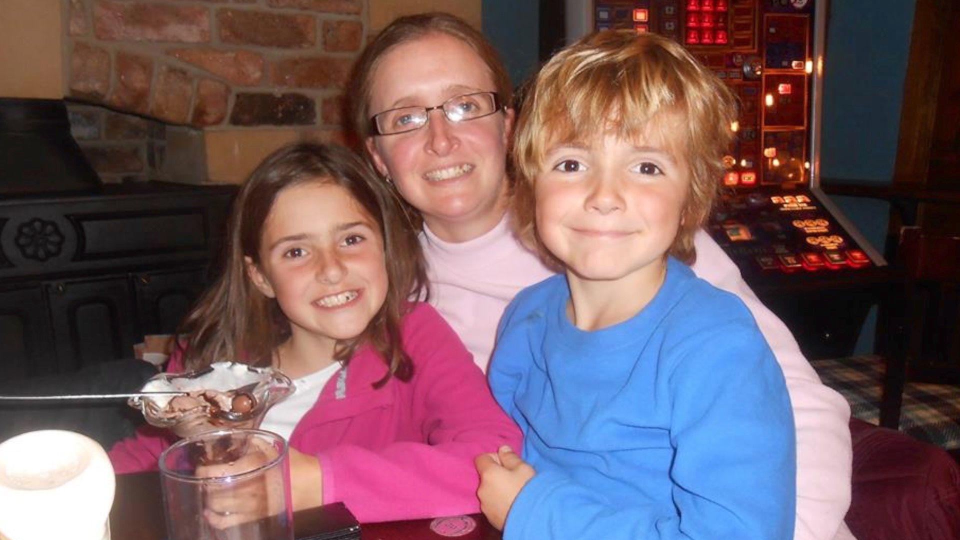 Carli Lansley in the centre of frame, with her daughter on the left and her son on the right, sitting at a table in a pub or restaurant. There is a brick fireplace and a gambling machine behind them. Carli is wearing a pink jumper and has dark-rimmed rectangular glasses, and has brown shoulder-length hair. Her daughter has dark brown shoulder-length hair, is wearing a white T-shirt with pink cardigan on top, and is holding an ice cream dessert. Carli's son is wearing a light blue round-neck jumper and has touseled blonde hair. All three are smiling at the camera.