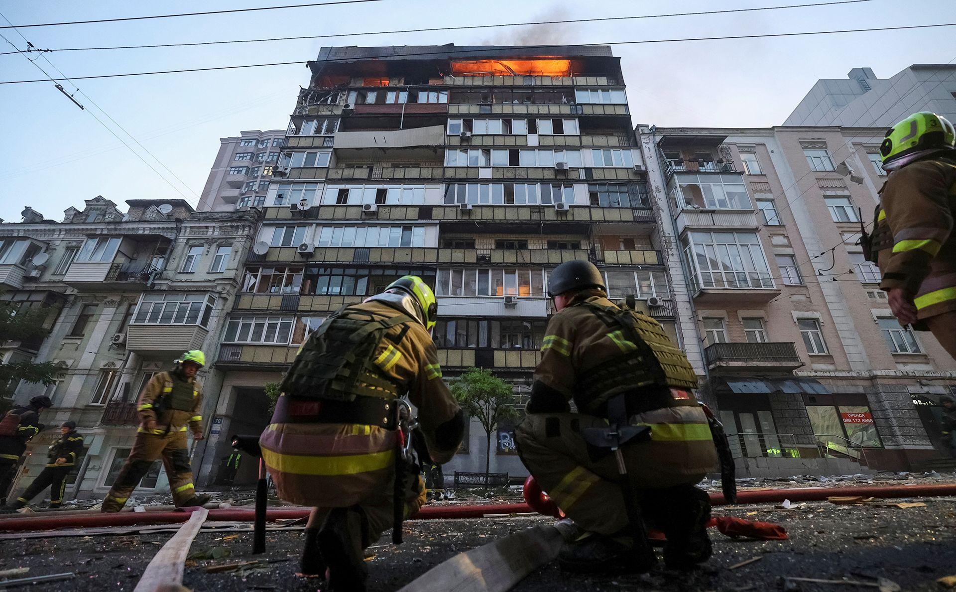 Firefighters crouch down on the street and look up at a block hit by a strike, with an active fire on the top floor against a blue sky in Kyiv on Thursday.