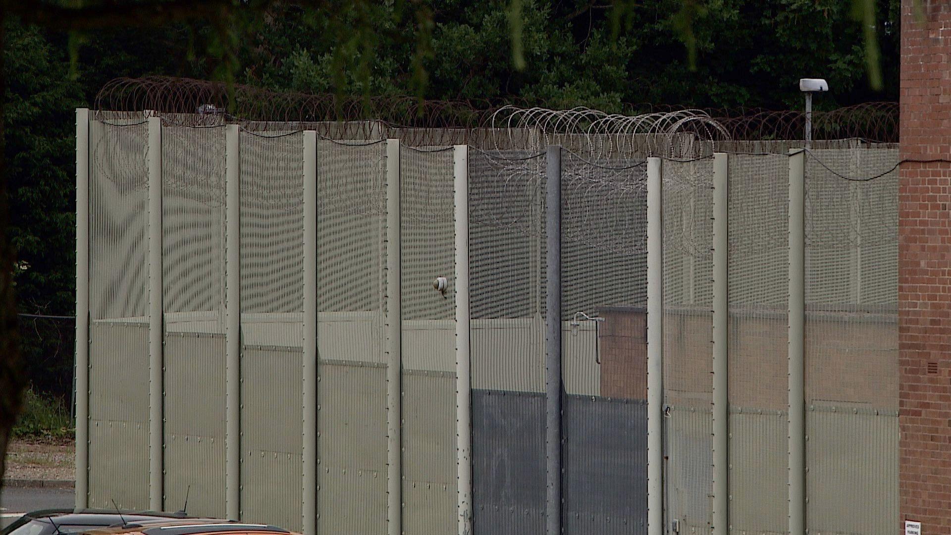 A barbed wire fence at Swinfen Hall prison. It is set against a backdrop of a grey sky. We can also see part of a high wall. 