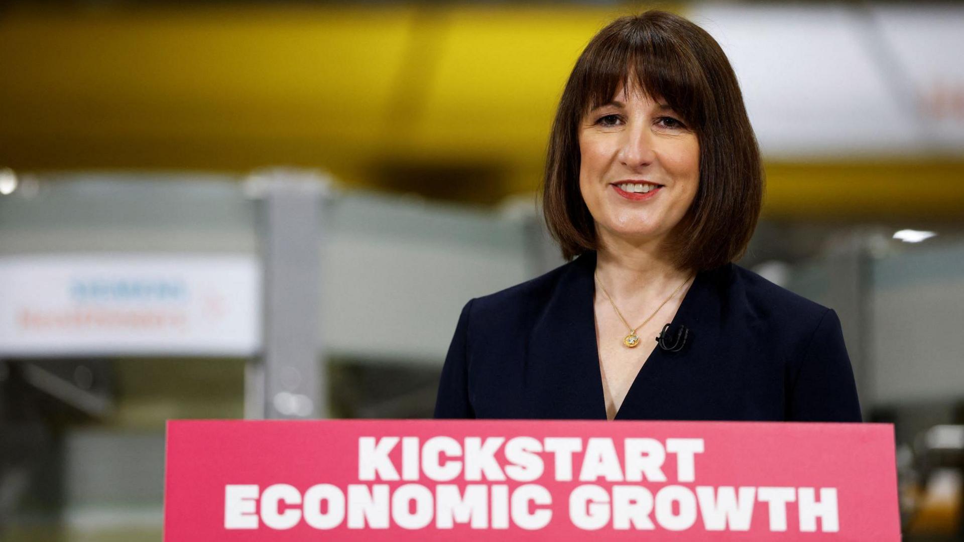 Rachel Reeves wearing a black jacket while standing in front of a red lectern, which has "kickstart economic growth" printed in white font on it.