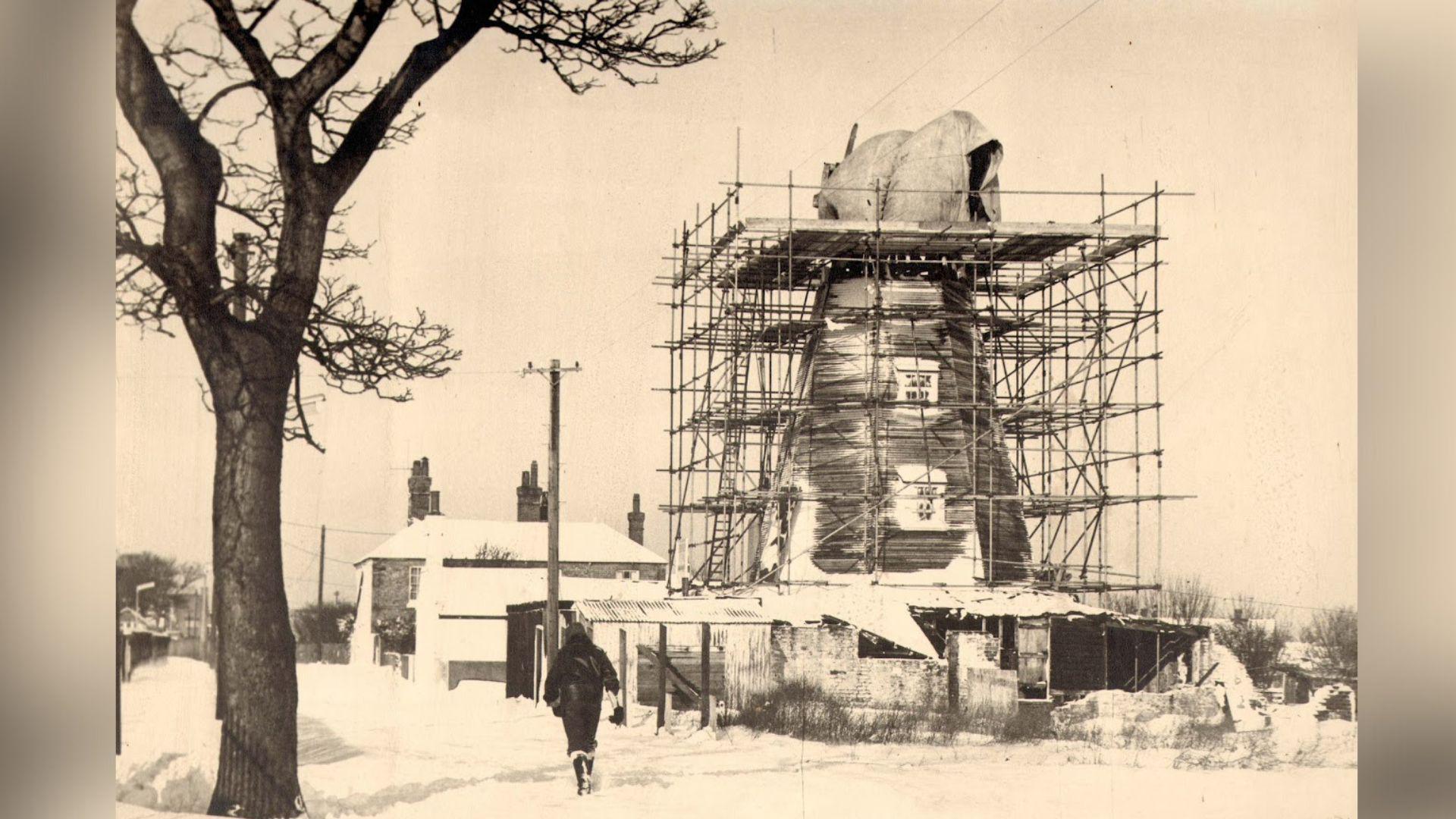A sepia photo of a windmill undergoing restoration. There are structural frameworks around the building.