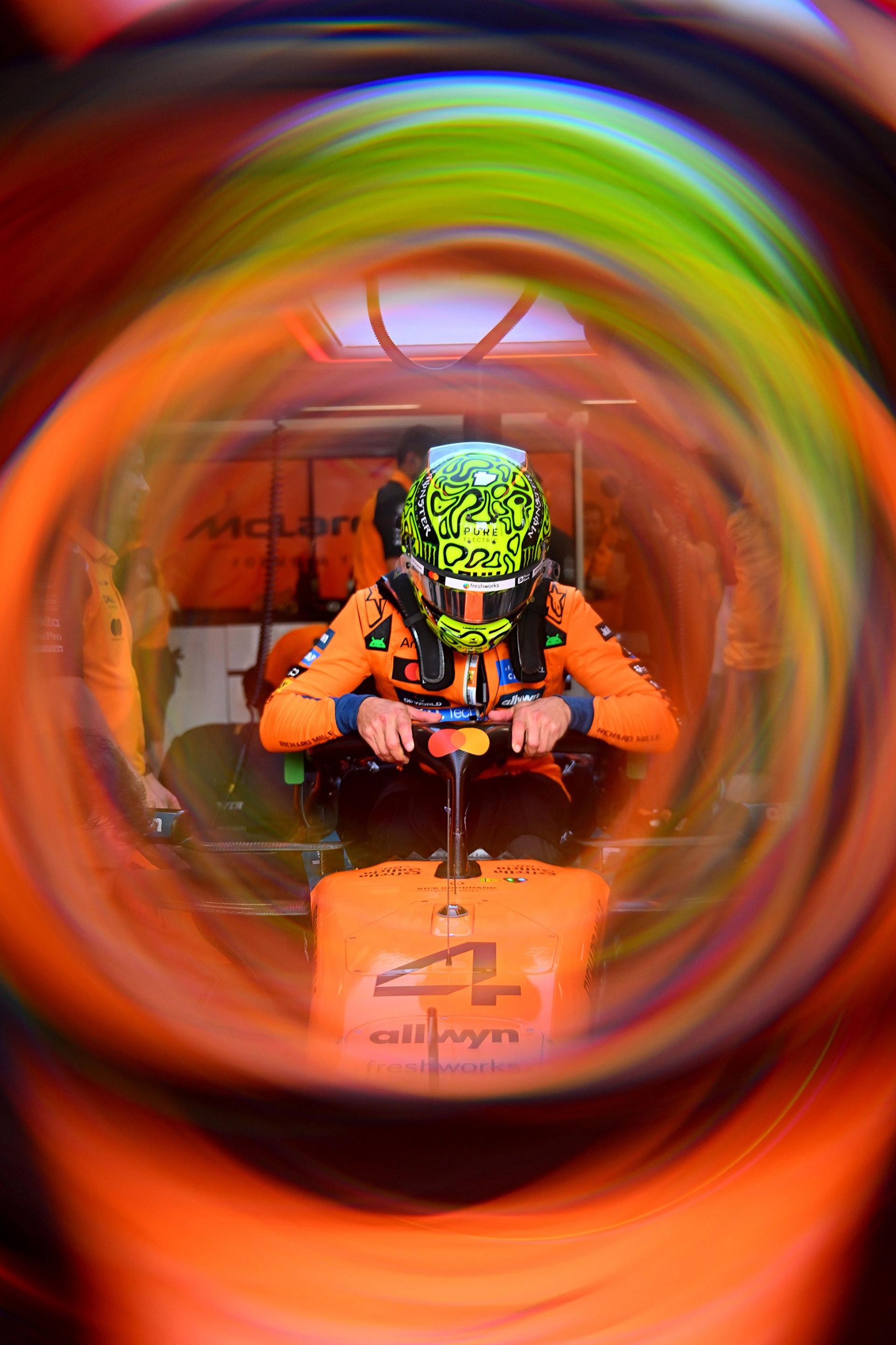 Driver wearing a bright green helmet with bold patterns seated in an orange McLaren Formula 1 car, viewed through a circular blur effect that creates a tunnel-like frame, emphasizing vibrant colors and the team branding in the background.