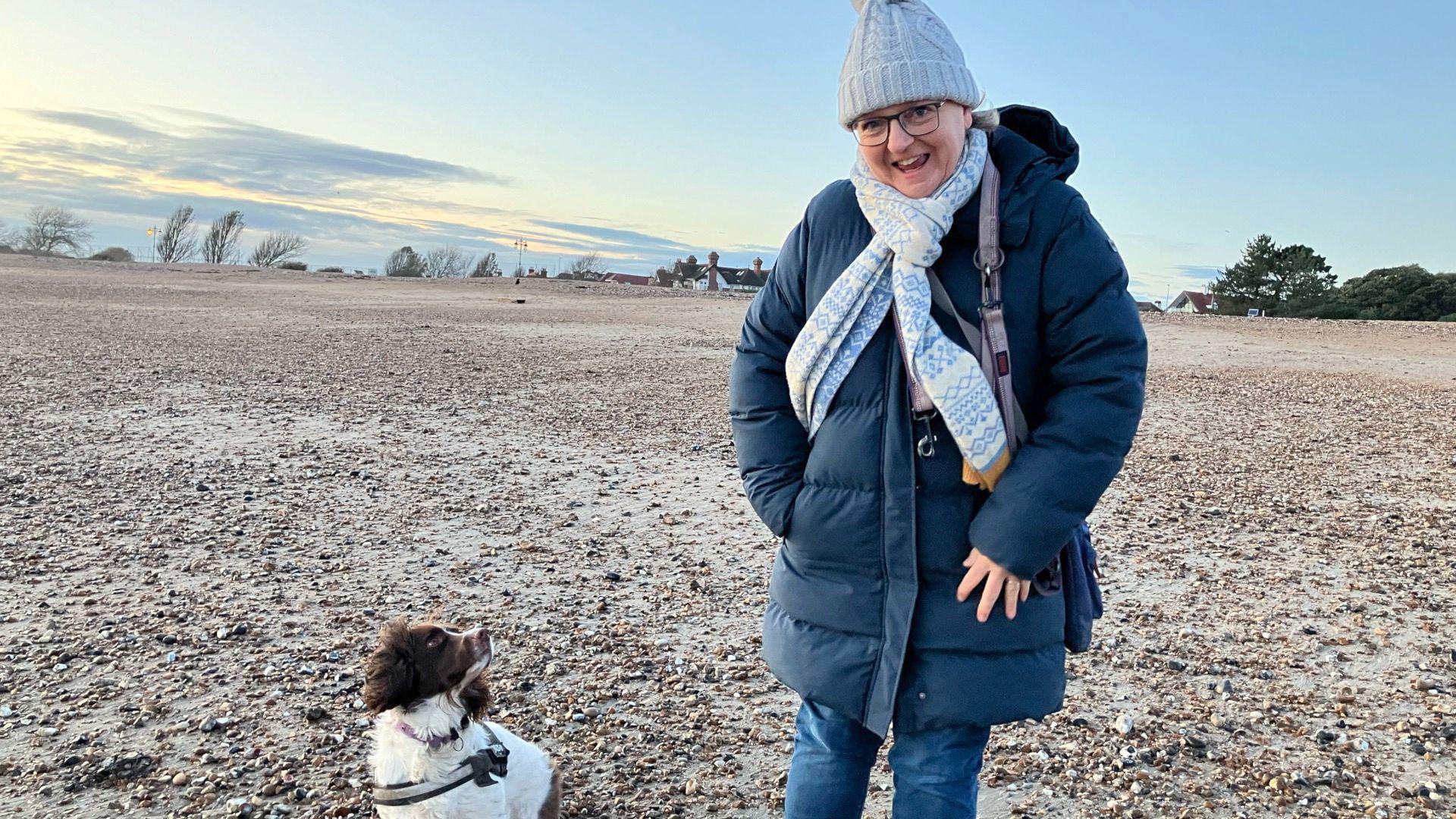 Sue Walker stands on Southsea beach, there are pebbles in the background and the tops of some trees can be seen further away. Sue is standing to the right of the frame, she's smiling at the camera and wearing a pale blue cable knit beanie with a fairisle print blue scarf and navy blue puffer duvet coat and blue denim jeans. She has a pruple lead draped over her should and her dog, Molly, a liver and white springer spaniel is sat to Sue's left, looking at her.